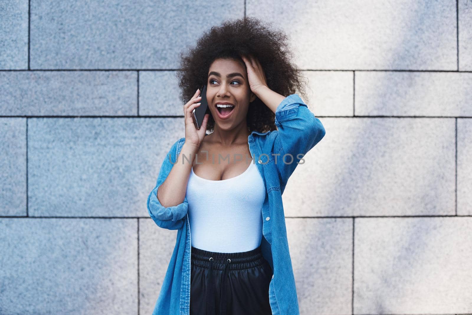 Young woman wearing jeans jacket free style on the street standing isolated on concrete wall talking on smartphone with friend touching head shouting surprised
