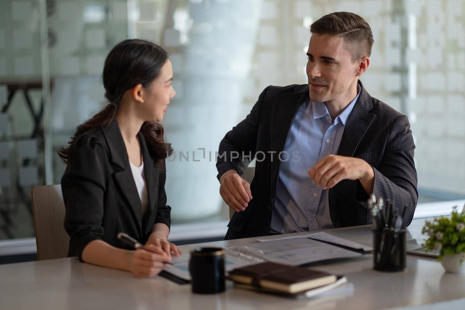 Multiethnic male caucasian mentor and female asian sitting at desk with laptop doing paperwork together discussing project financial report. Corporate business collaboration concept.