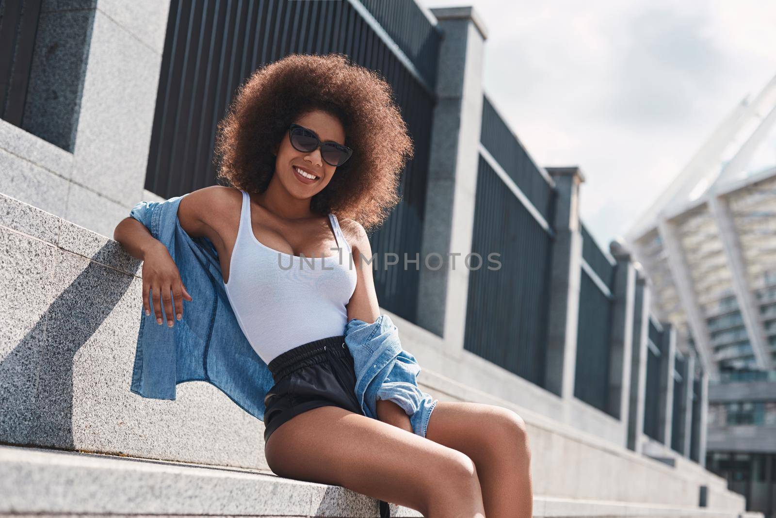 Young woman wearing sunglasses free style on the street sitting on concrete stairs leaning back relaxed looking camera smiling cheerful