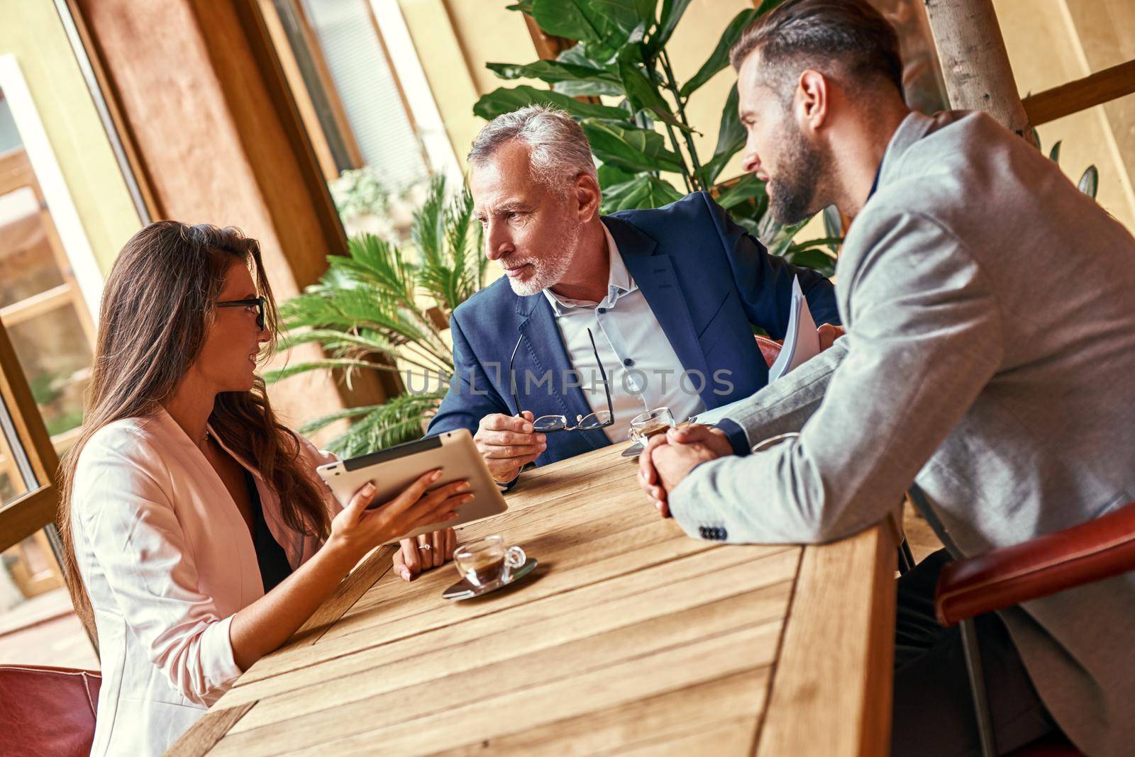 Business lunch. Three people in the restaurant sitting at table discussing project woman holding digital tablet explaining ideas looking at senior man smiling cheerful by friendsstock