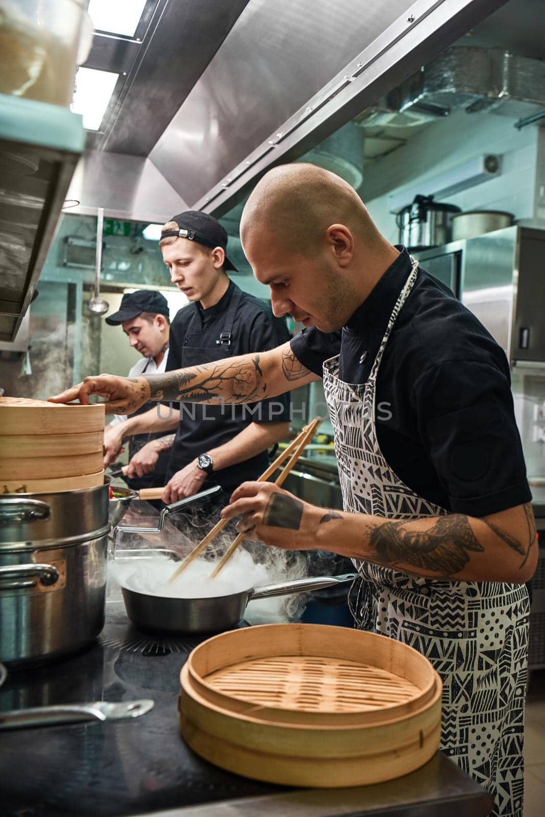 Full concentration. Young bald chef in apron with tattoos on his arms preparing dish with his two assistants in a restaurant kitchen. Cooking process