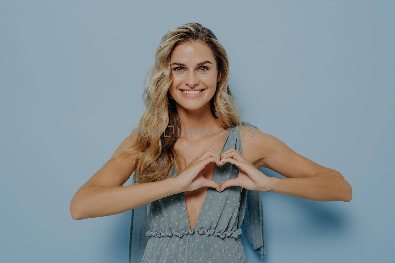 Happy lovely young female with blonde long hair showing love symbol with her hands cupped in heart shape, caucasian woman in elegant evening dress, sending tender passion, radiating positive emotions