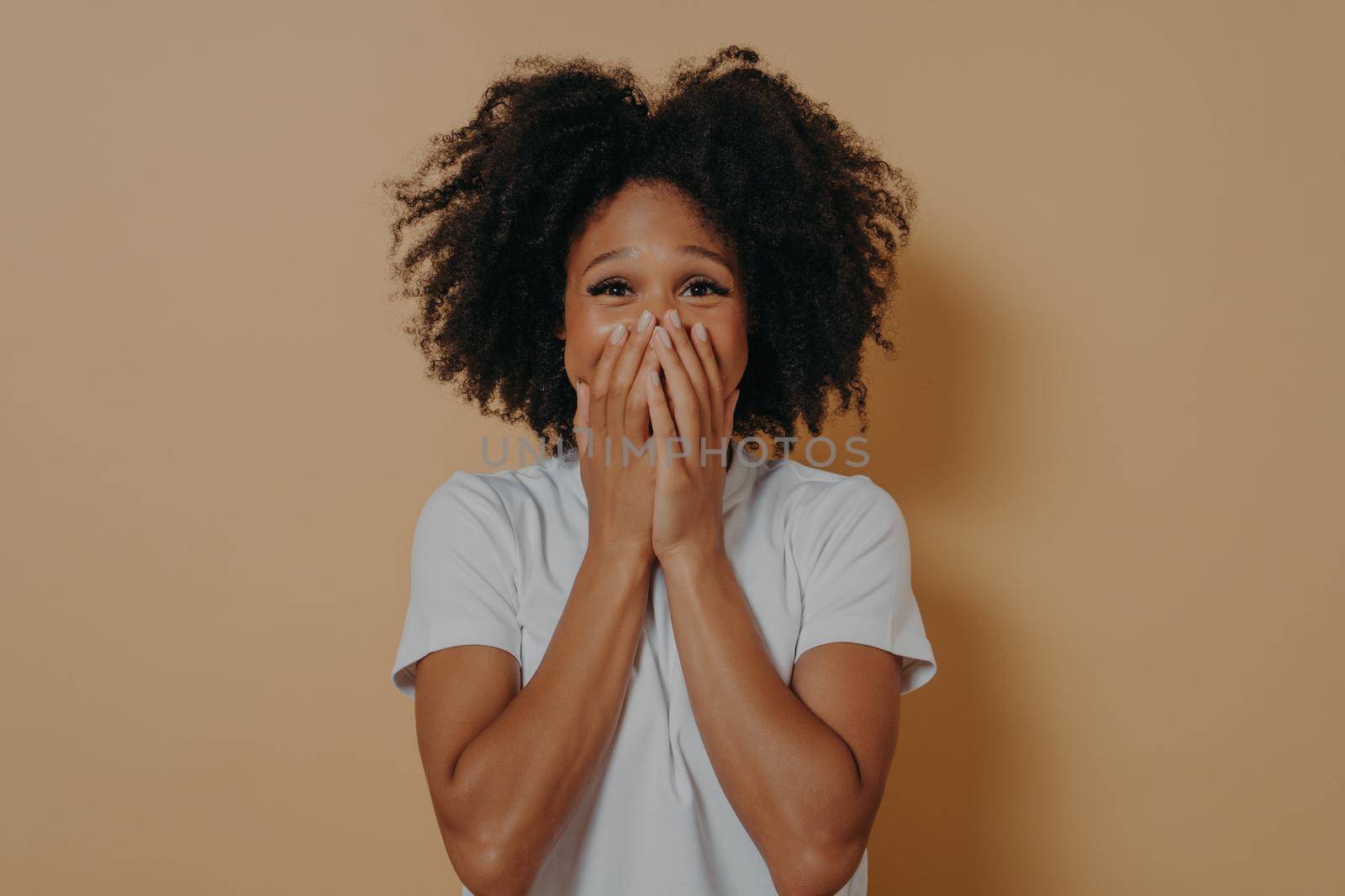 Surprised excited dark skinned woman in white tshirt covering her mouth with hands isolated on pastel beige background with copy space. Positive women emotions and body language concept