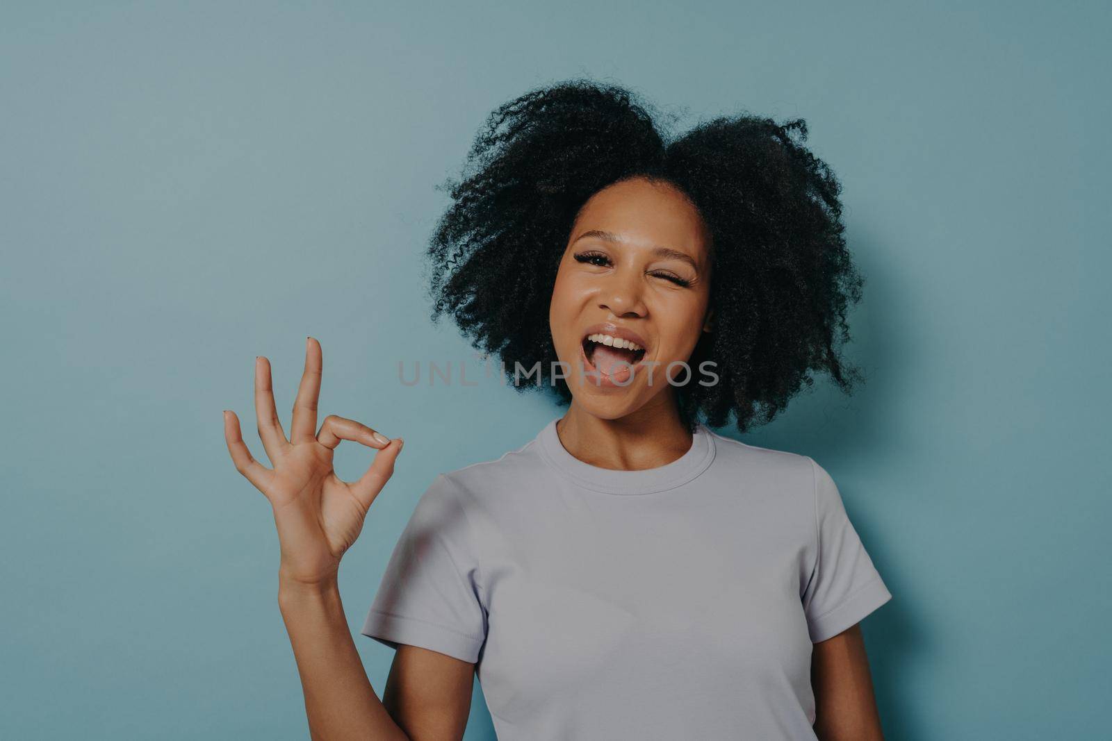 Pretty girl of mixed race appearance showing ok gesture with fingers and winks, showing that everything is great. Attractive African female in good mood posing in studio, looking friendly at camera