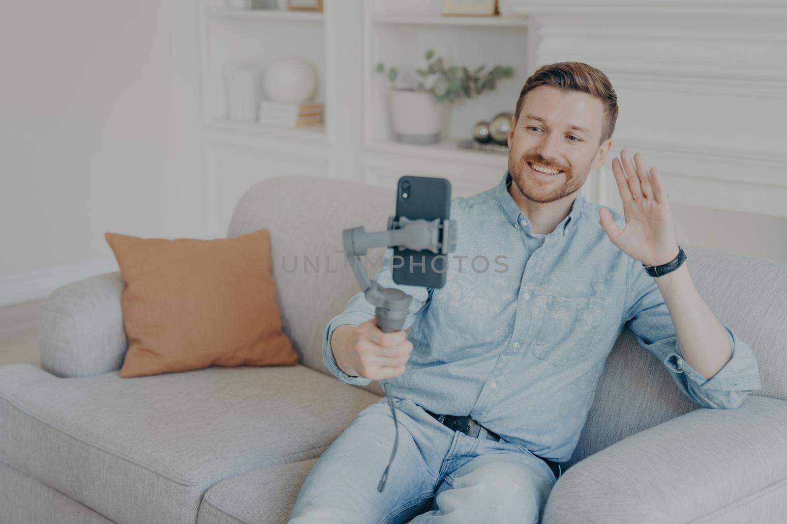 Young man enjoying video chat with friends , looking at phone screen and waving with hand, cheerful bearded man holding smartphone with gimbal stabilizer handheld while sitting on sofa in living room
