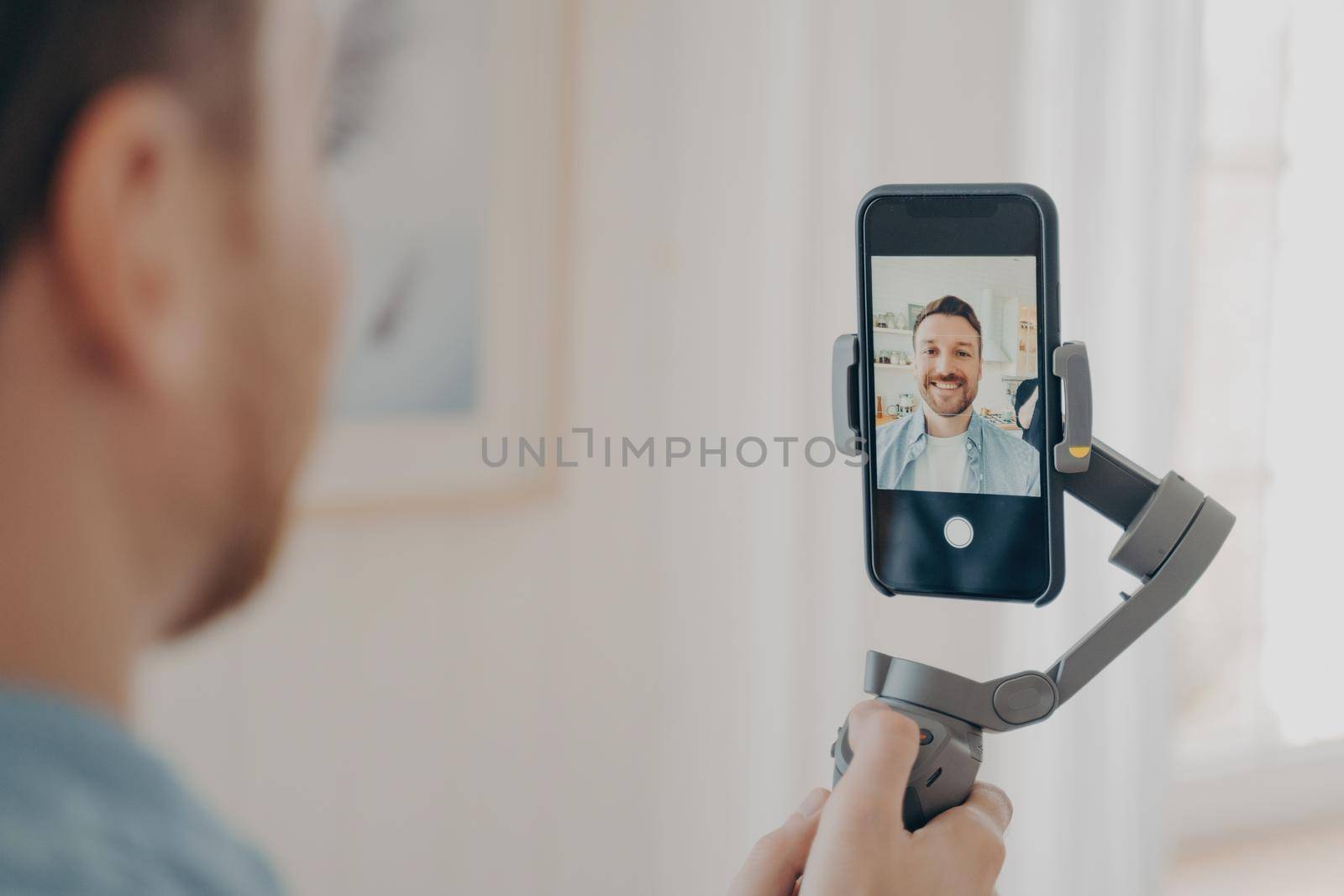Handsome young man vlogging or recording video on smartphone with great handheld gimbal stabilizer, standing in living room at home background. Vlog and video blogging concept