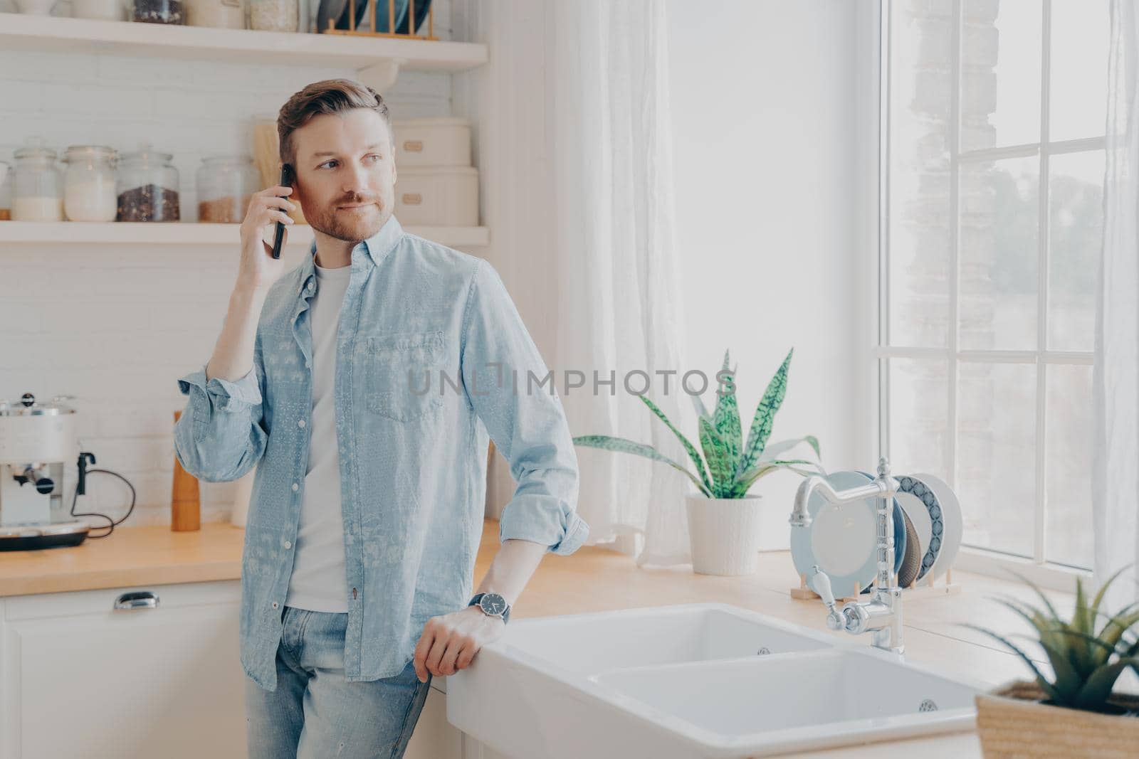 Young male in casual jeans shirt speaking on cellphone with work partner and looking away while standing by wooden table top in modern kitchen interior. Freelance and distance work concept