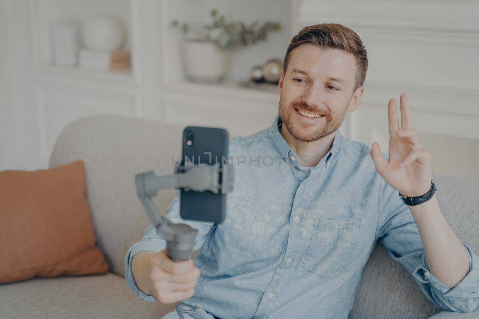 Young man talking with his friend through online video chat using telephone attached to gimbal, waving hello with hand while smiling, happy after not seeing each other for long time