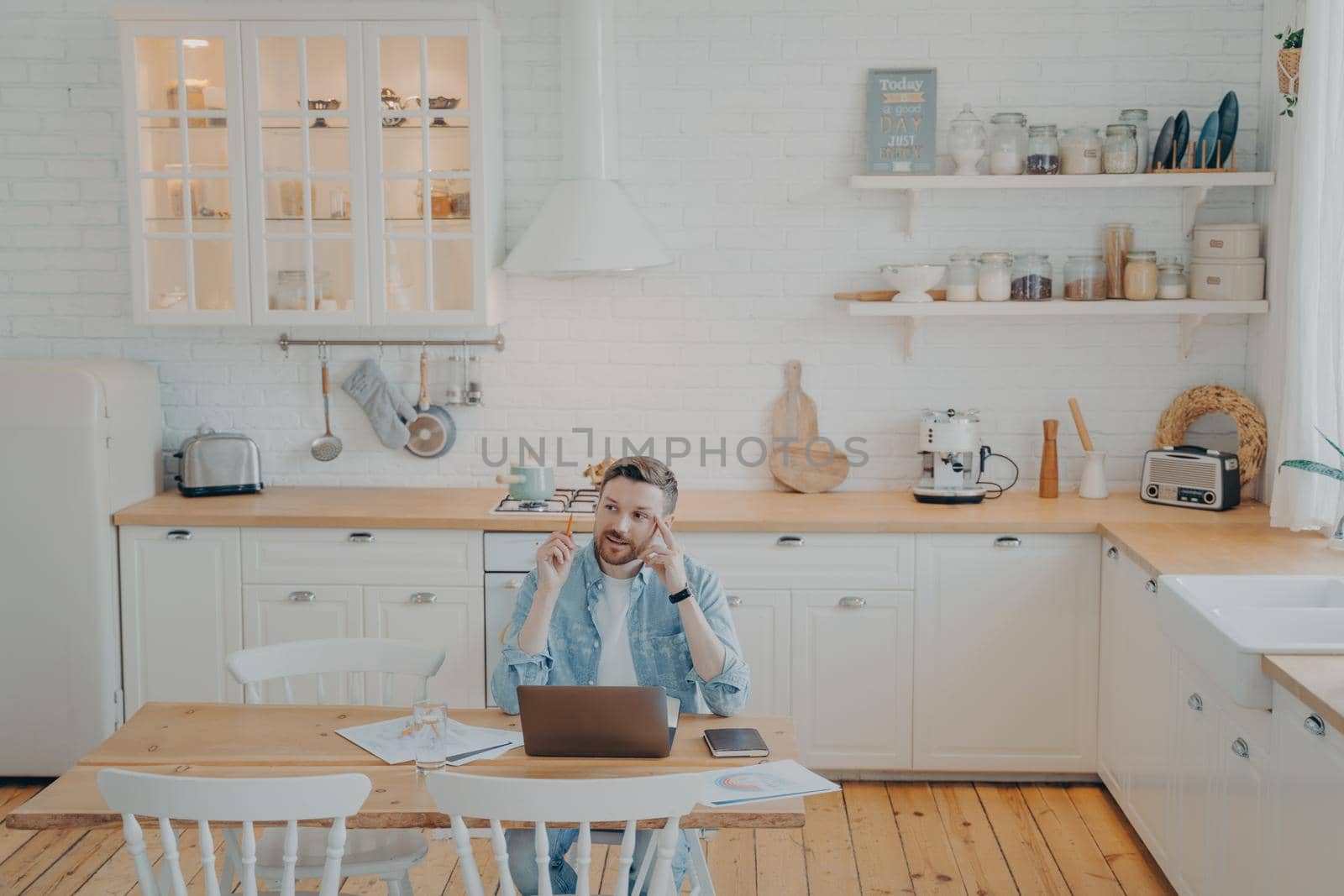 Young office worker working remotely from home with computer while sitting by kitchen table by vkstock