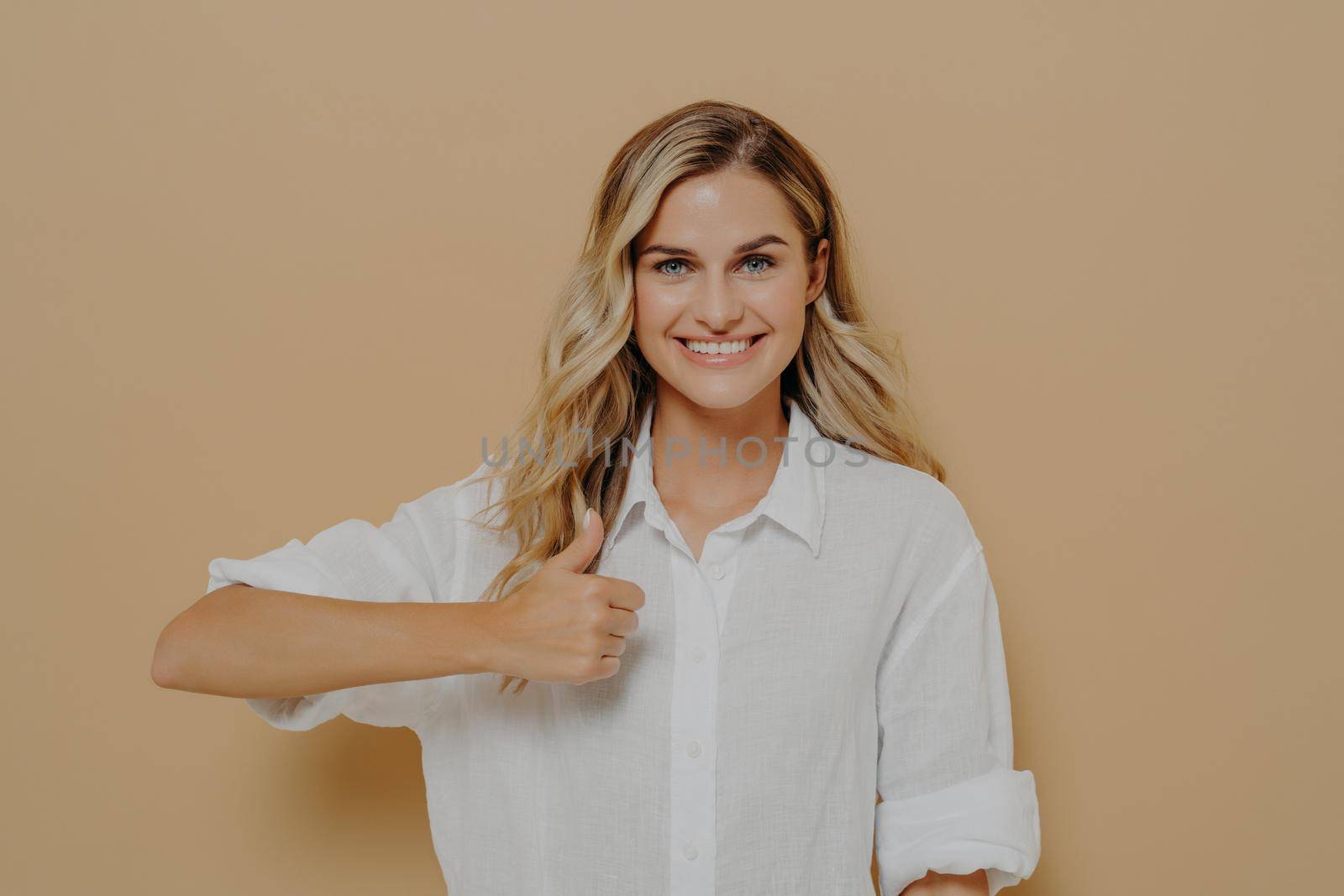 Achieving goals. Portrait of positive young woman with broad smile showing thumb up gesture with hand while posing in studio against beige wall, looking at camera with happy expression. Body language