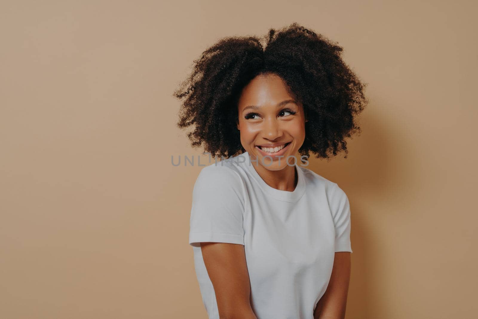 Cute african woman with toothy smile looking aside with shyness, isolated over beige background by vkstock