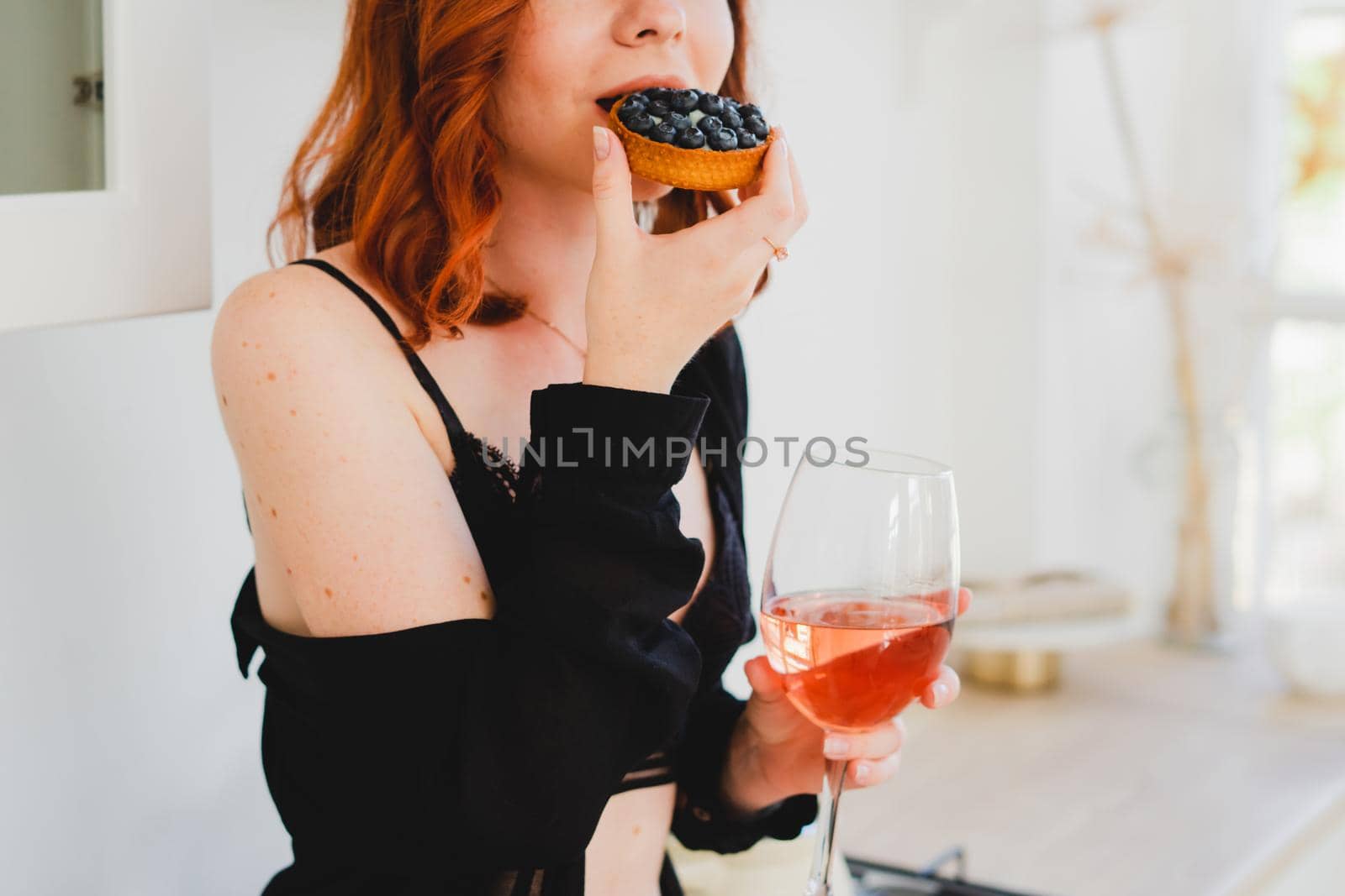 The girl is sitting eating a cake and drinking wine. A young girl in a black negligee is sitting in the kitchen eating a cake and drinking wine. Bright kitchen. Sexy girl is resting.