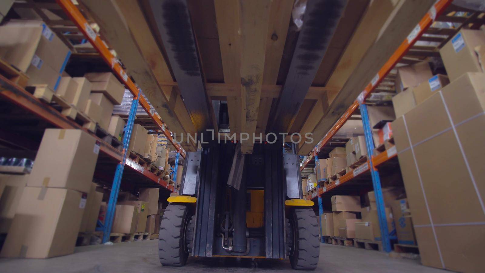 Worker drives forklift trucks with cardboard boxes in warehouse. Man rides along metal racks in the storehouse