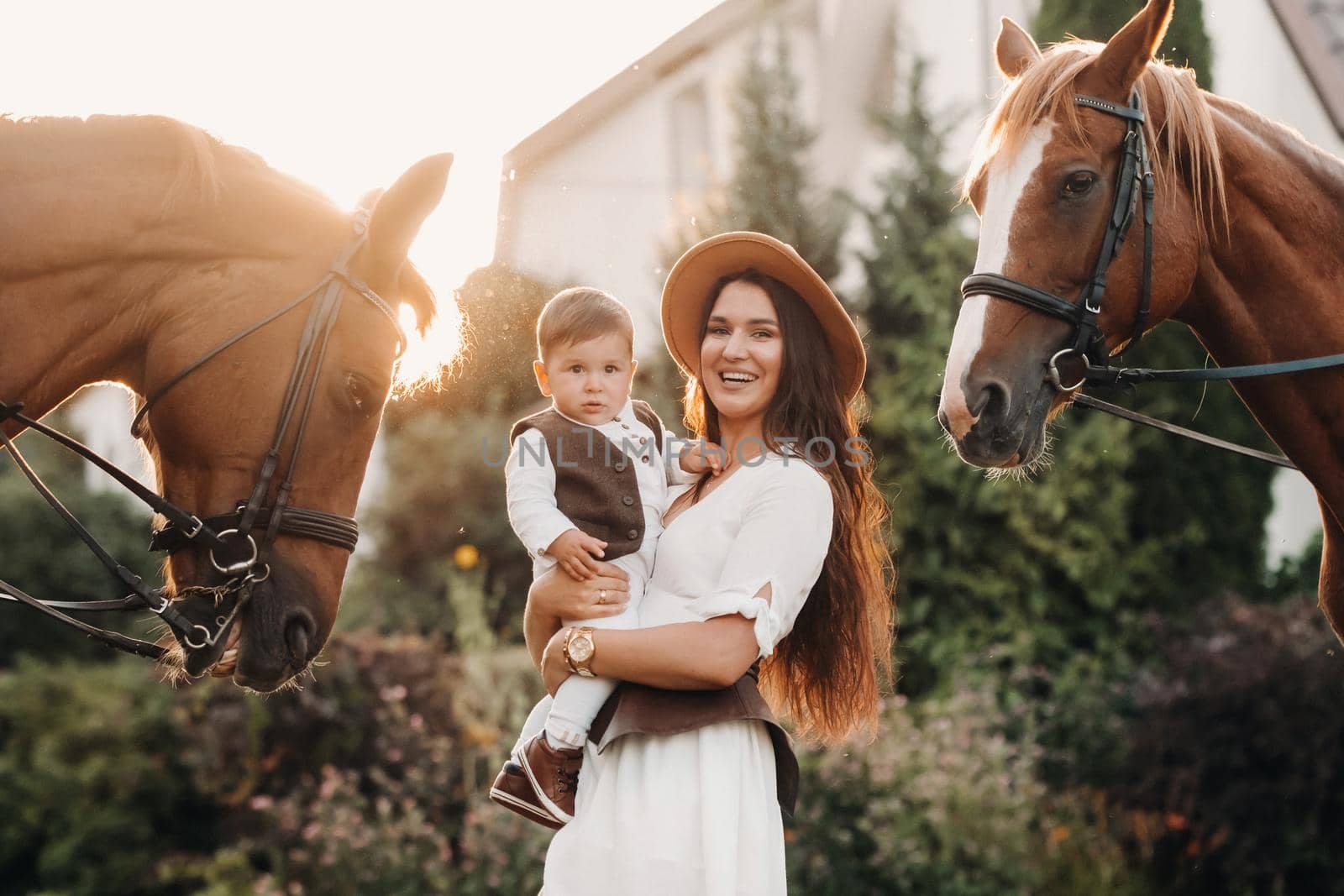 a mother in a hat with her son in her arms stands next to two beautiful horses in nature. a family with a child is photographed with horses.