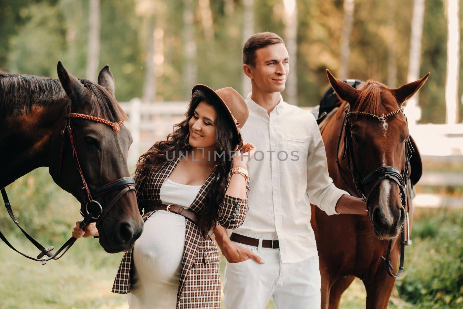 a pregnant girl in a hat and her husband in white clothes stand next to horses in the forest in nature.Stylish pregnant woman with a man with horses.Family. by Lobachad