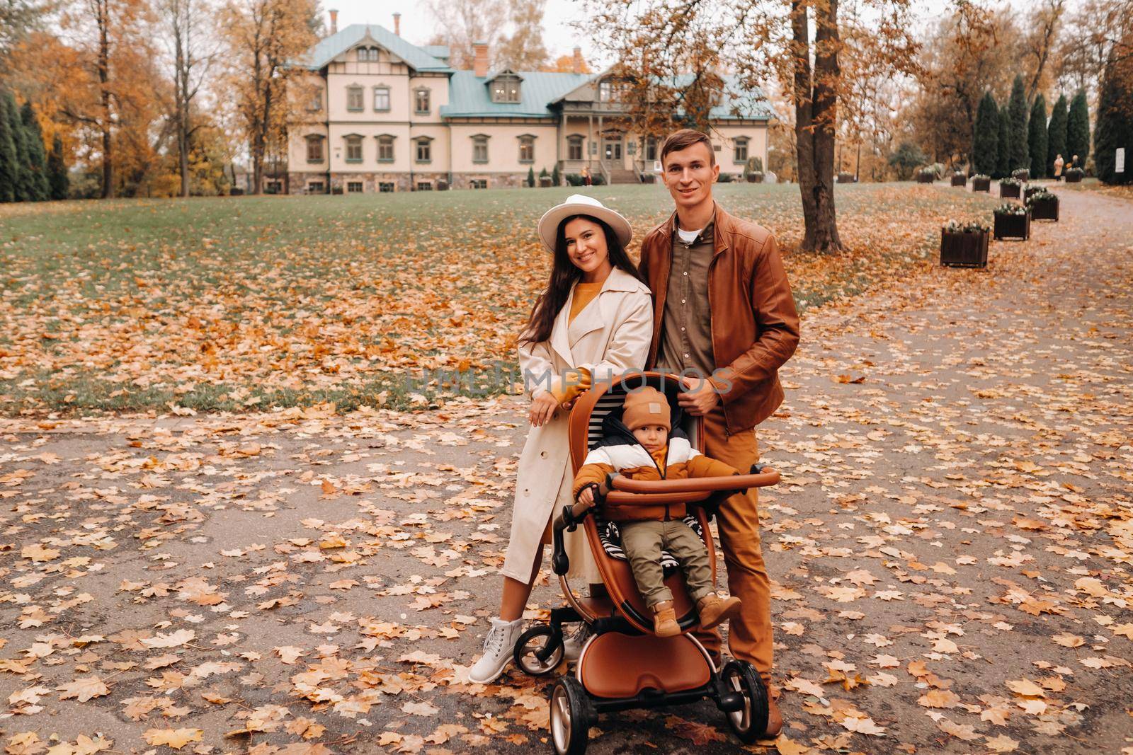 Father and mother on a walk with a child in a stroller in the autumn Park on the background of the estate. A family walks through the Golden autumn nature Park.