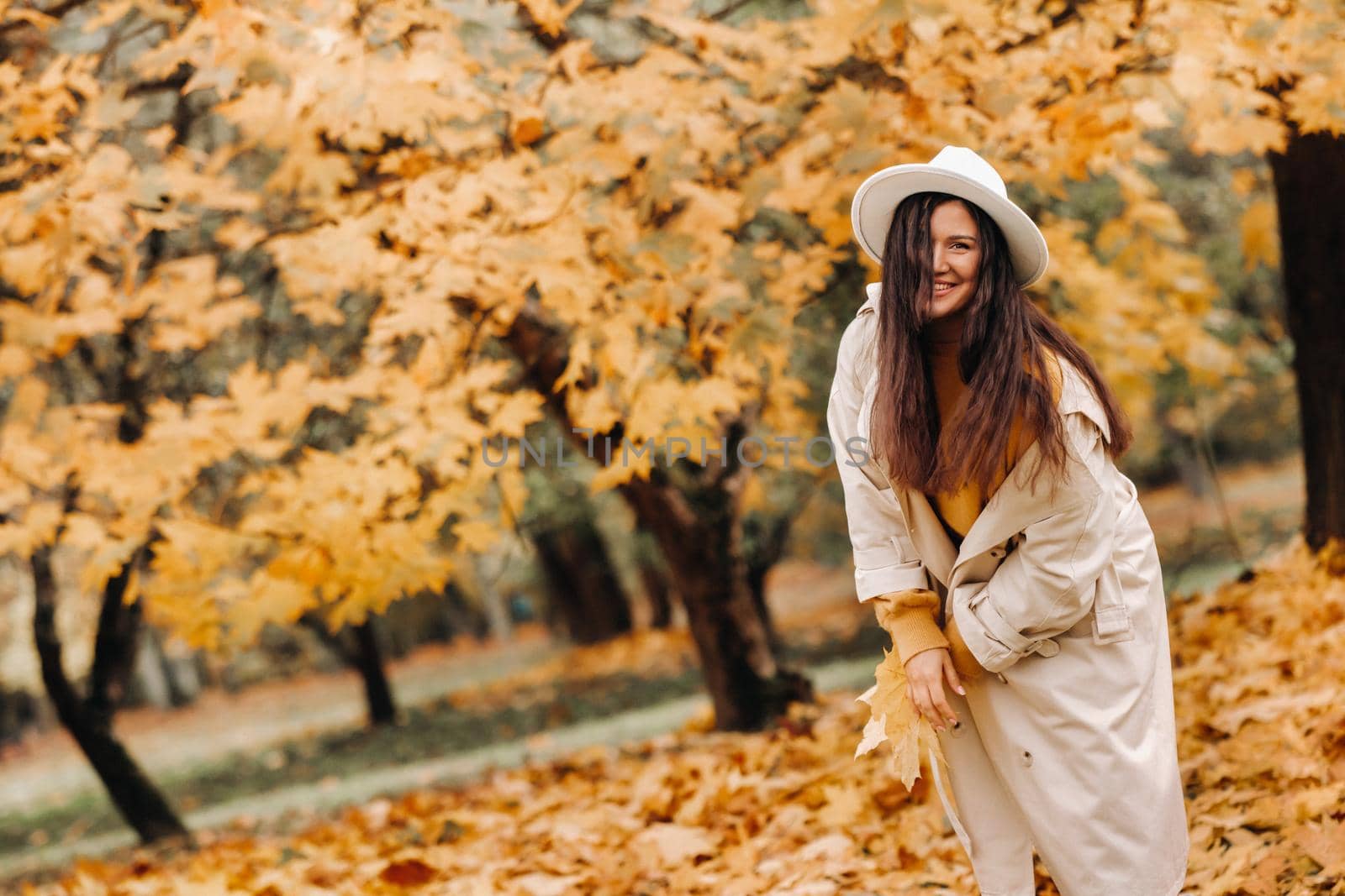 a cheerful girl in a white coat and hat smiles in an autumn Park.portrait of a smiling woman in Golden autumn. by Lobachad