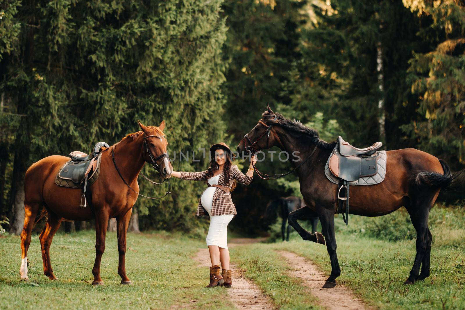 pregnant girl with a big belly in a hat next to horses in the forest in nature.Stylish pregnant woman in the brown dress with the horses