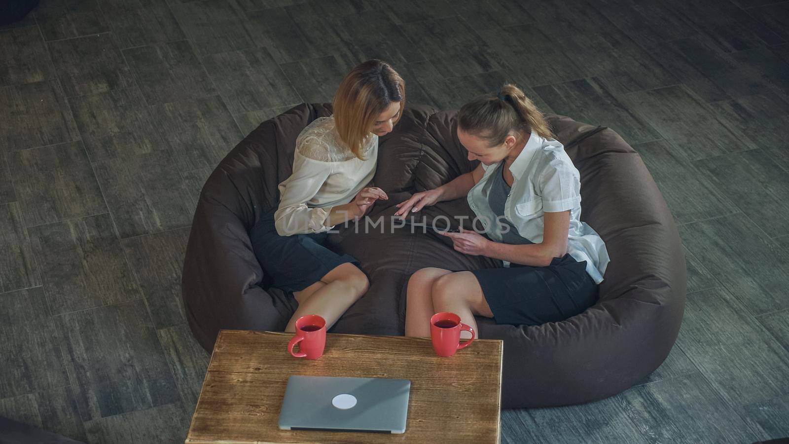 Two women at the cafe watching photos on digital tablet and laughing. On the wooden coffee table red cups with tea