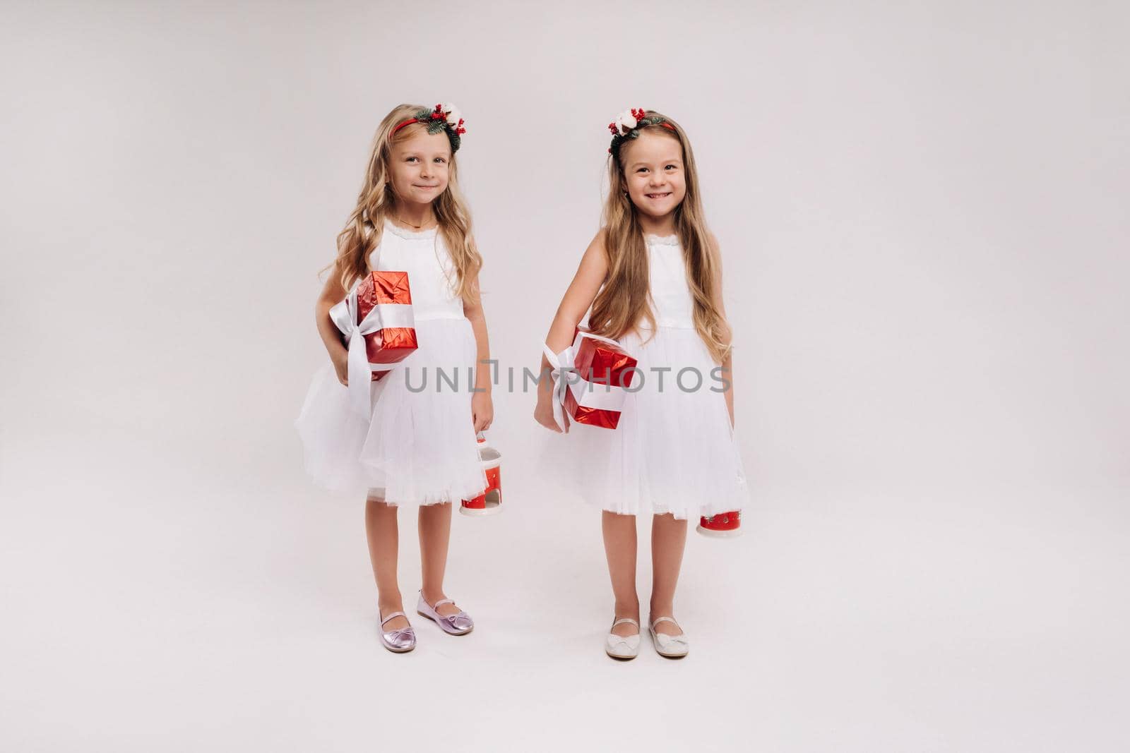 two little girls in white dresses with Christmas gifts on a white background smile.