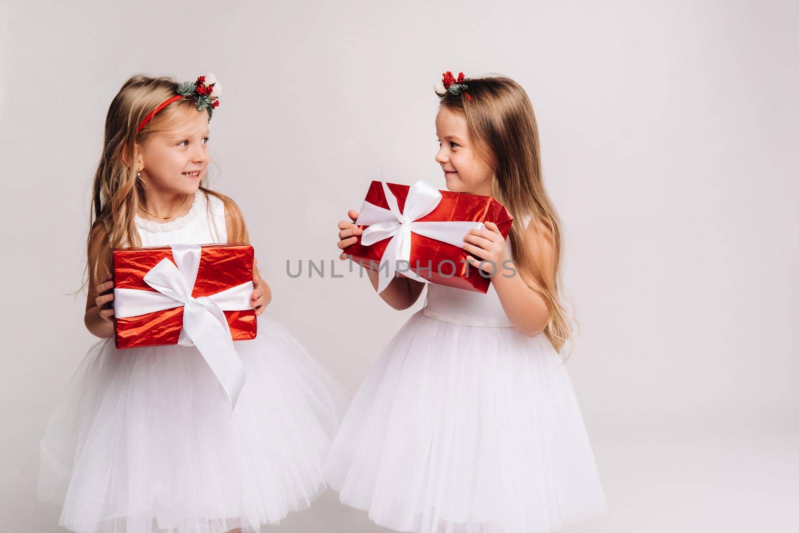 two little girls in white dresses with Christmas gifts on a white background smile by Lobachad