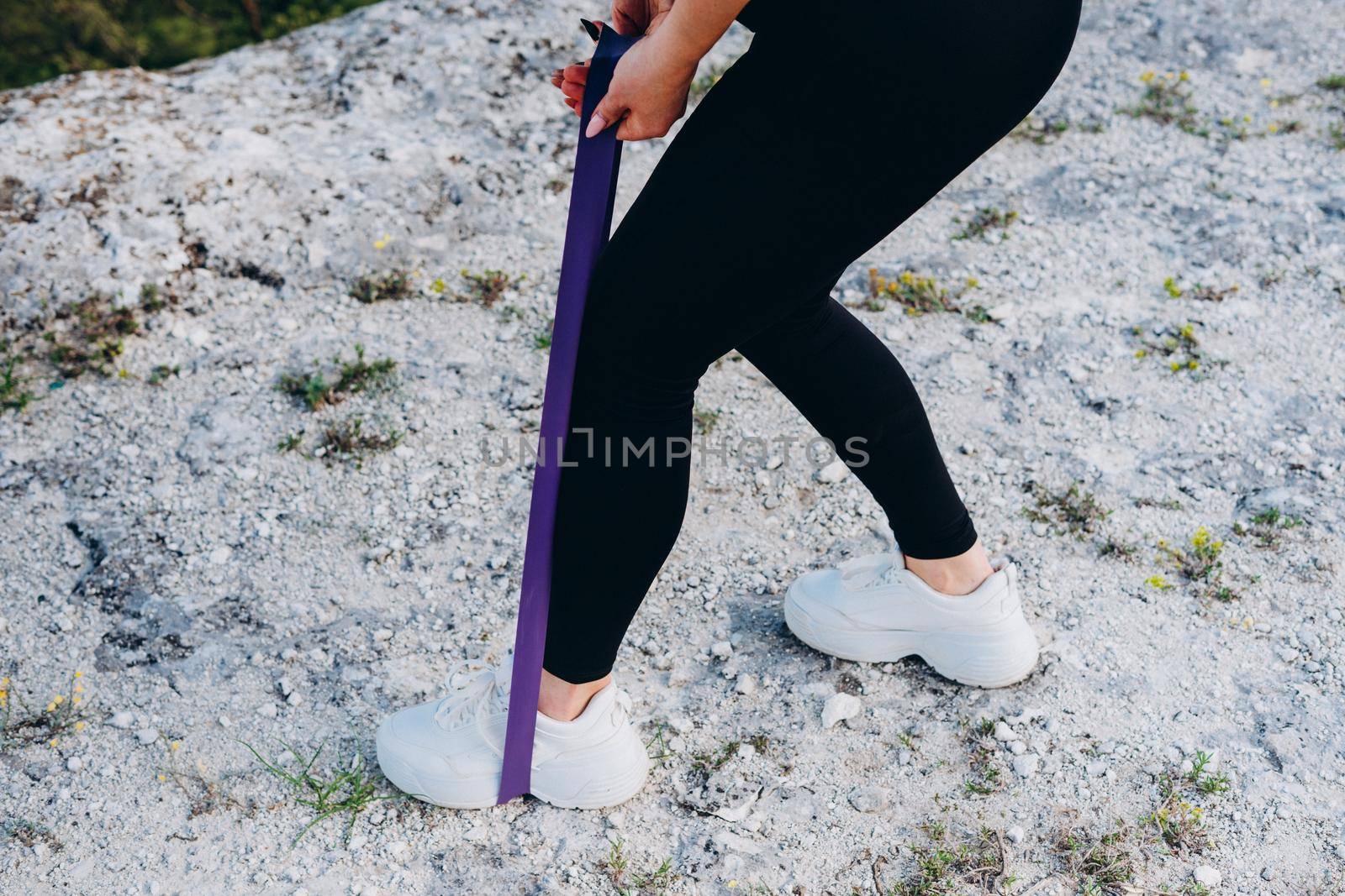 The girl does an exercise for the legs with the help of a rubber band. A beautiful athlete in the mountains.