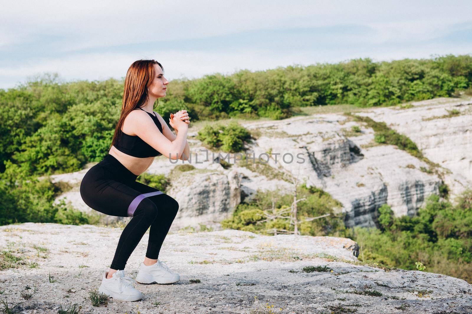 The girl is engaged in sports in nature. Purple rubber band for stretching and swaping muscles of hands and feet. A beautiful athlete in the mountains.