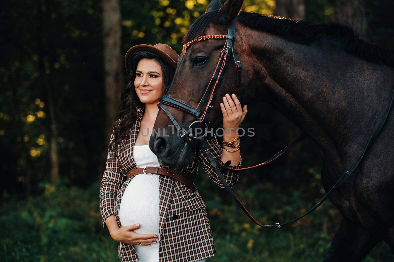 pregnant girl with a big belly in a hat next to horses in the forest in nature.Stylish girl in white clothes and a brown jacket. by Lobachad