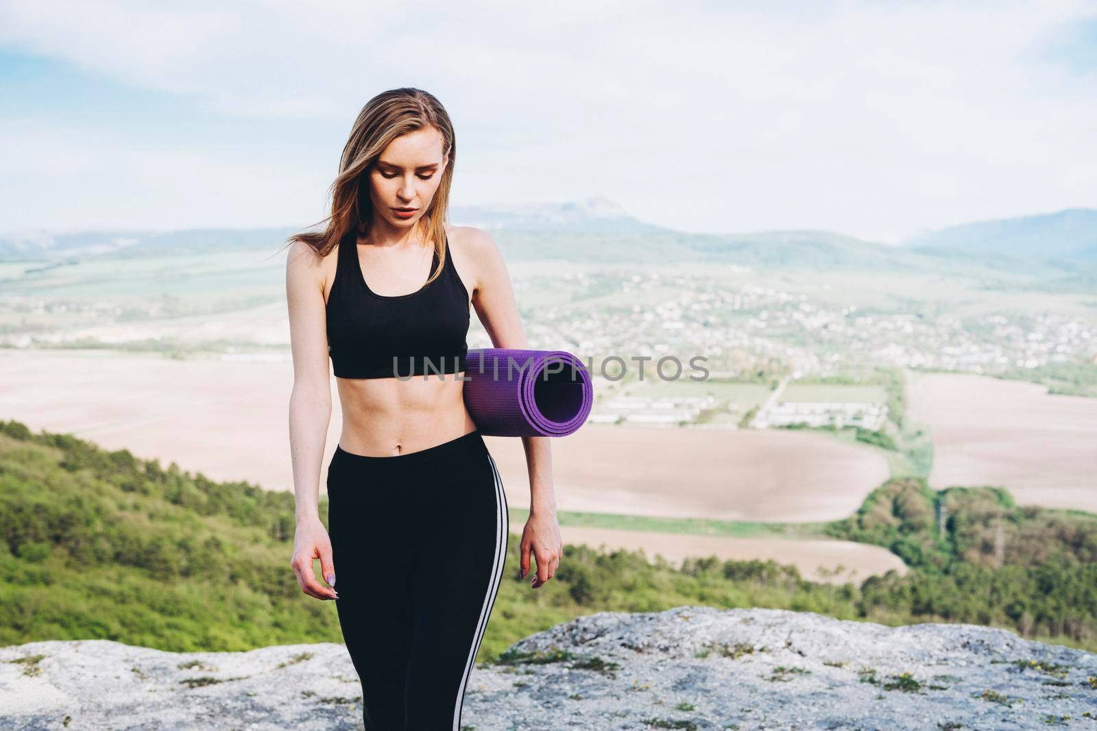 A girl athlete with a purple yoga mat under her armpit. A beautiful athlete in the mountains.