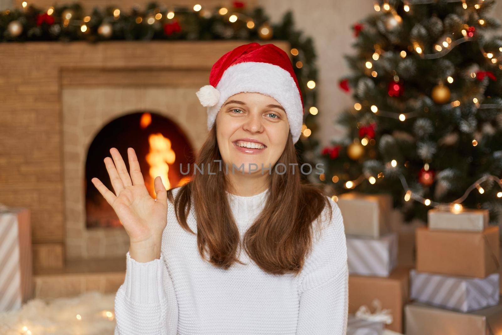 Young woman smiling happily and cheerfully, waving hand, welcoming and greeting somebody or saying goodbye, wearing santa hat and sweater, posing near christmas tree, fireplace and boxes with gifts.