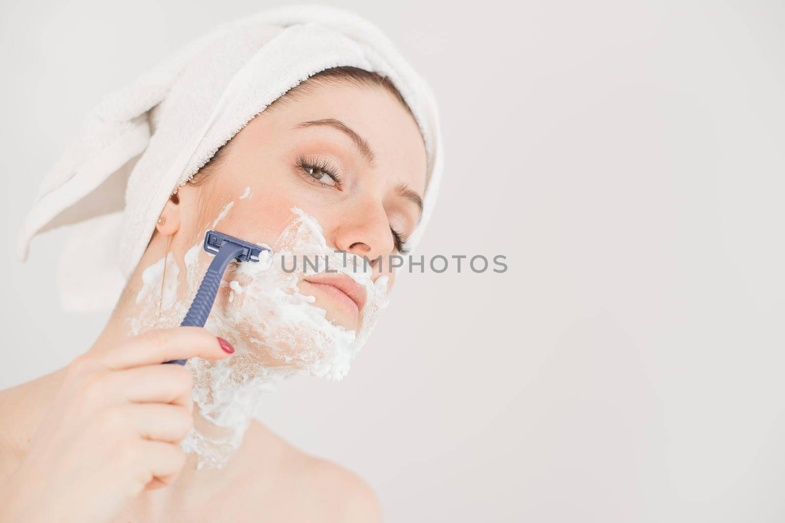Cheerful caucasian woman with a towel on her head and shaving foam on her face holds a razor on a white background by mrwed54