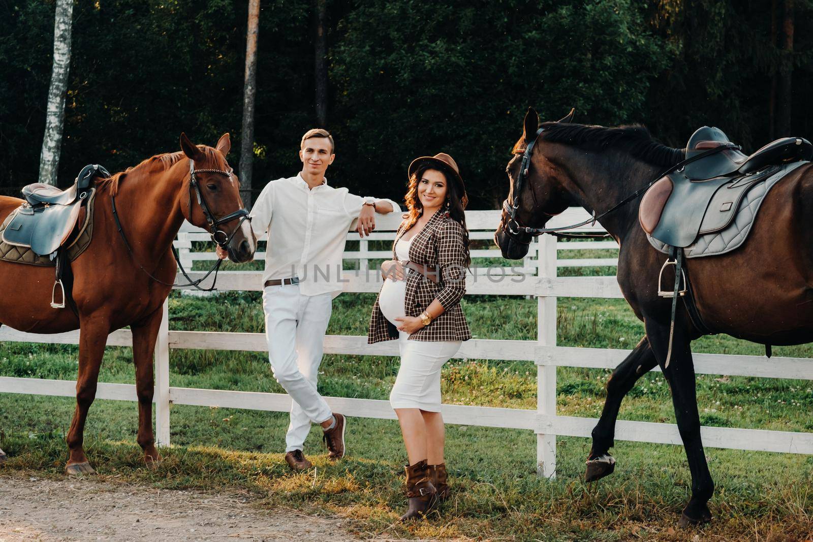 a pregnant girl in a hat and a man in white clothes stand next to horses near a white fence.Stylish pregnant woman with a man with horses.Married couple