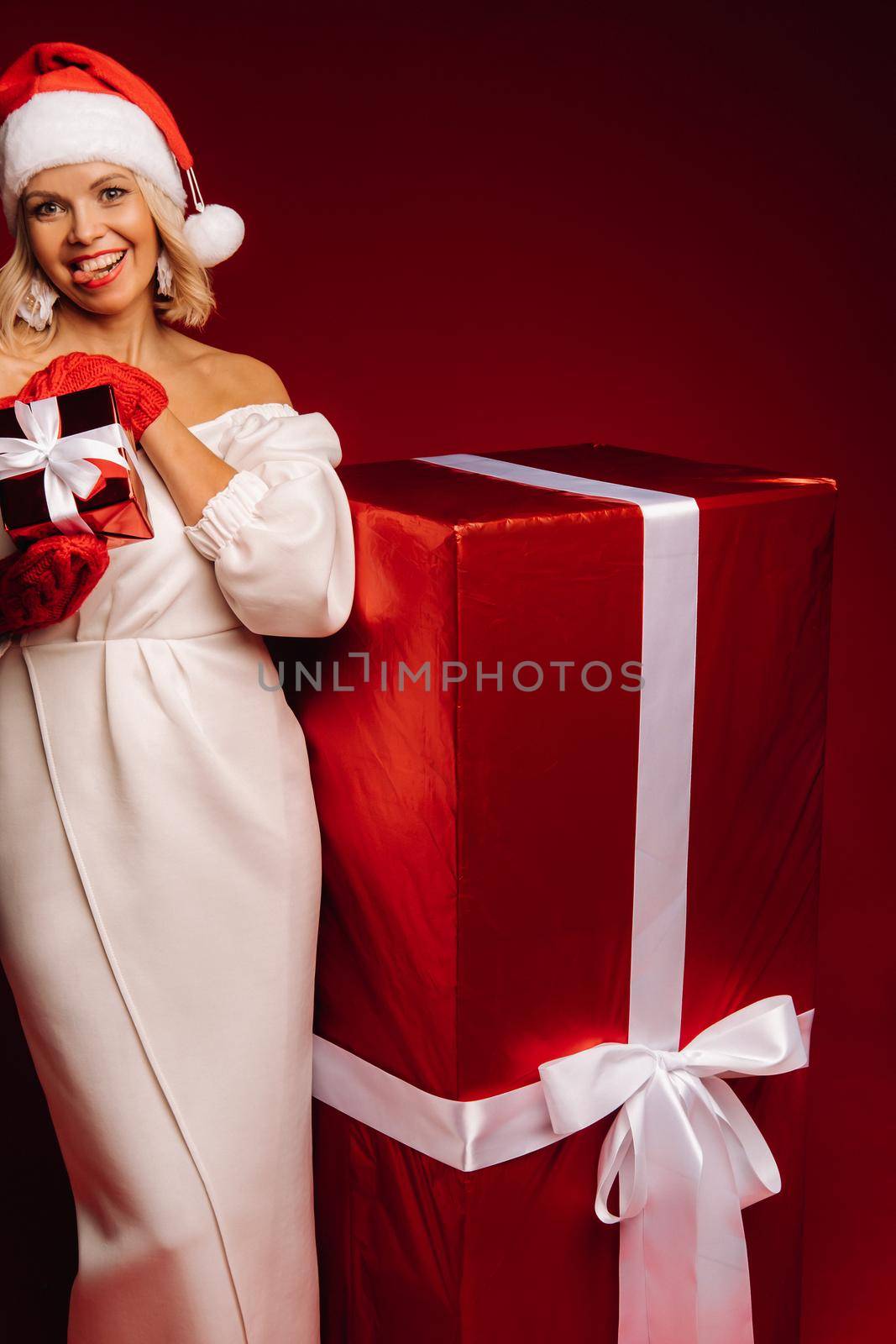 portrait of a smiling girl in a white dress and Santa hat with a Christmas gift on a red background.