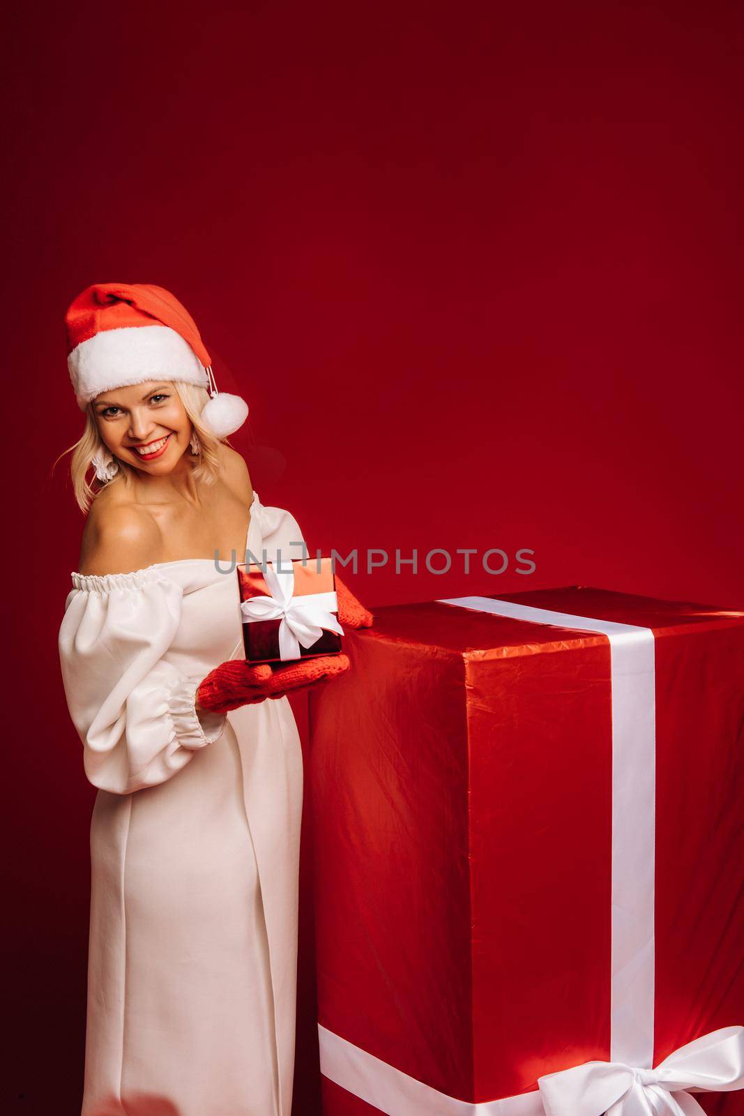 portrait of a smiling girl in a white dress and Santa hat with a Christmas gift on a red background by Lobachad