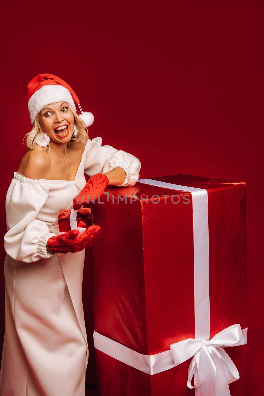 portrait of a smiling girl in a white dress and Santa hat with a Christmas gift on a red background.