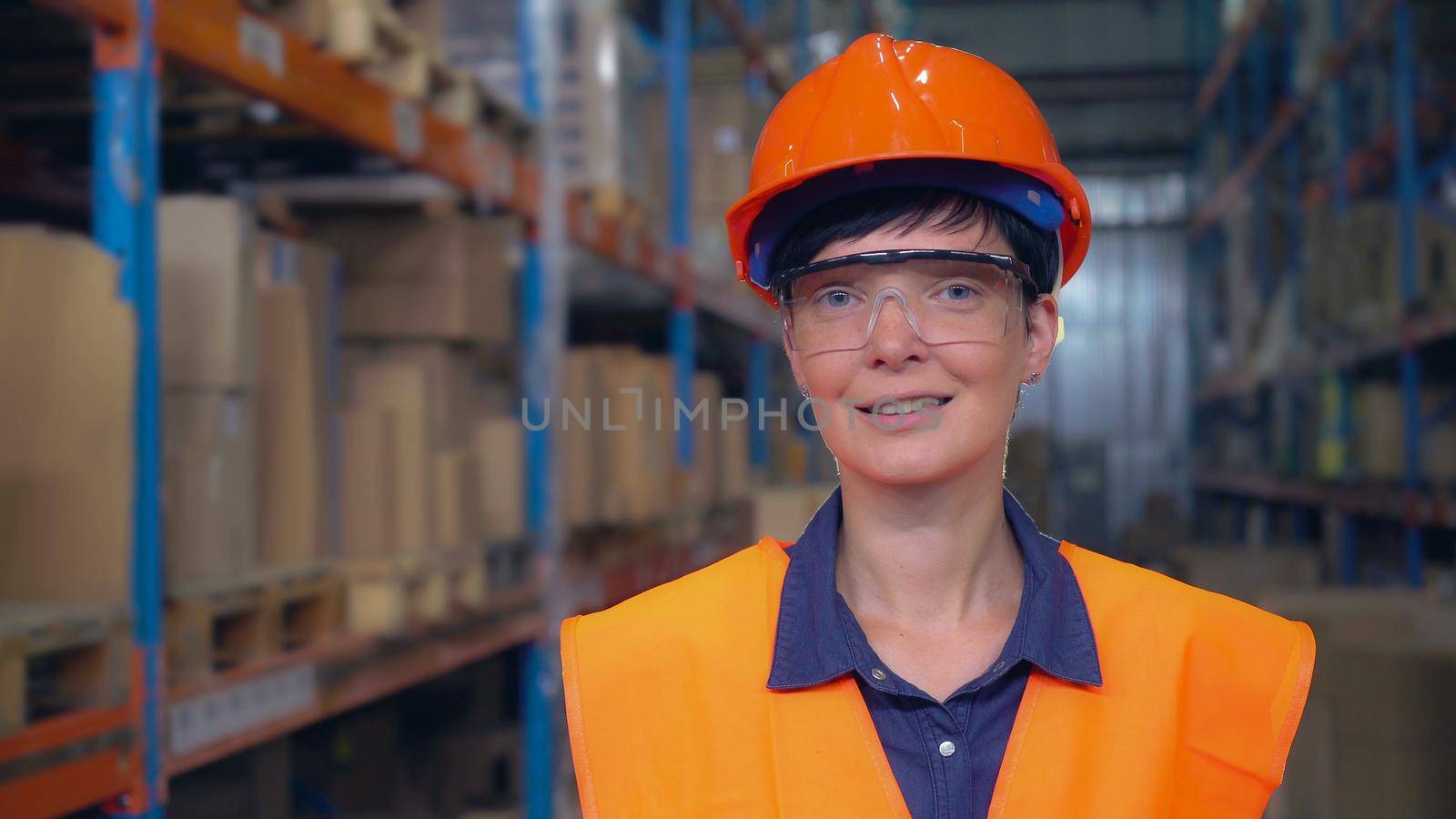 Woman worker in warehouse posing looking at the camera. Employee wearing in orange hard hat and uniform. Young female standing near metal racks with boxes.