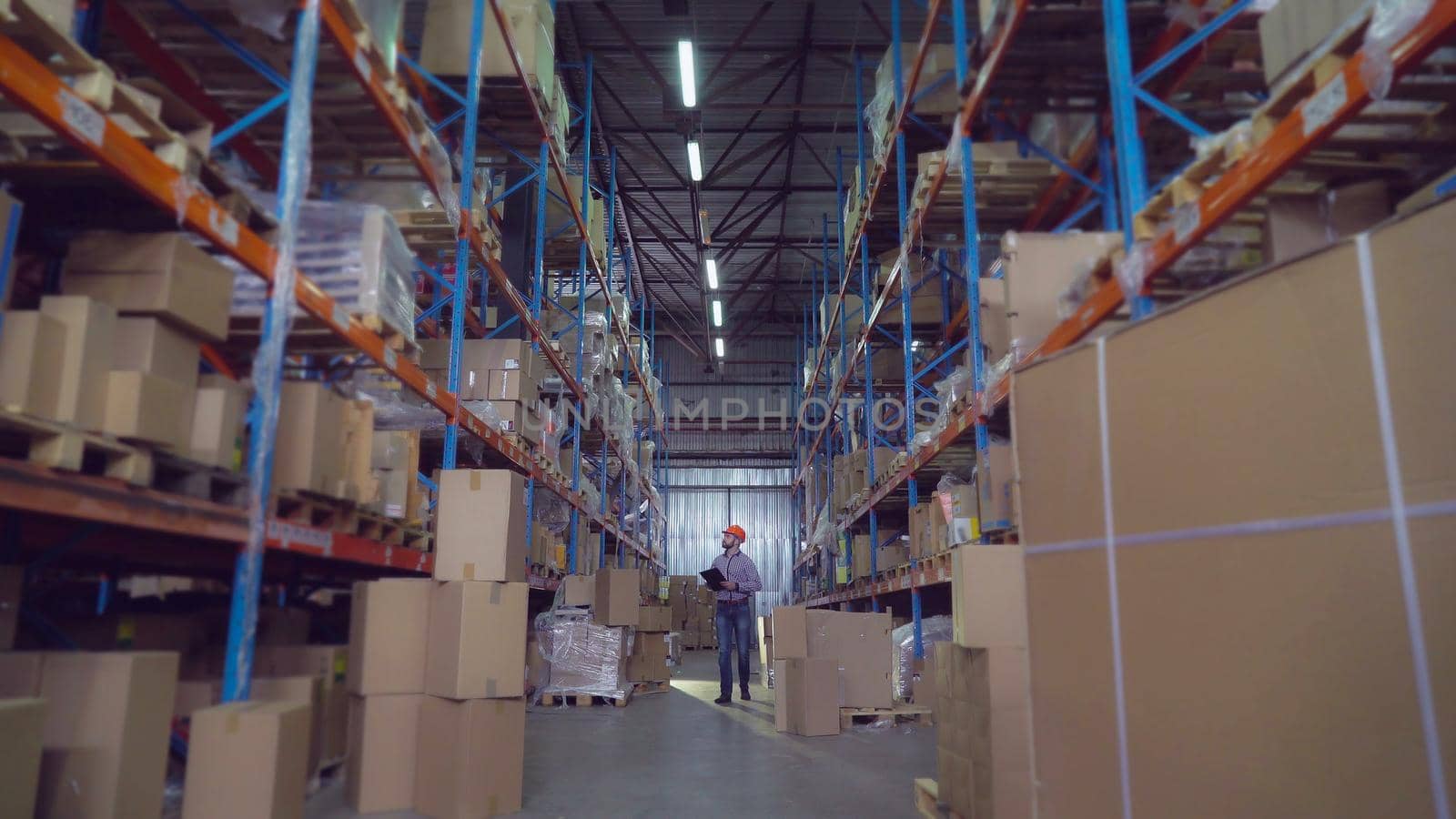 Man workers warehouse going along racks. Manager wearing hard hat writing notes in paper documents checking stock. Young employee with beard wearing casual shirt and jeans.