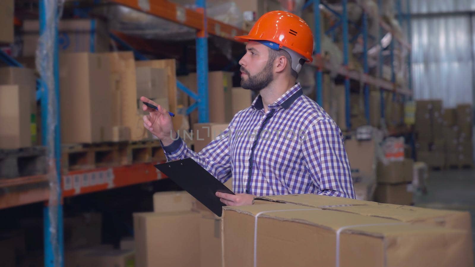 Handsome young worker preparing delivery in logistic center. Man wearing casual shirt and hard hat writing some notes or filling form.