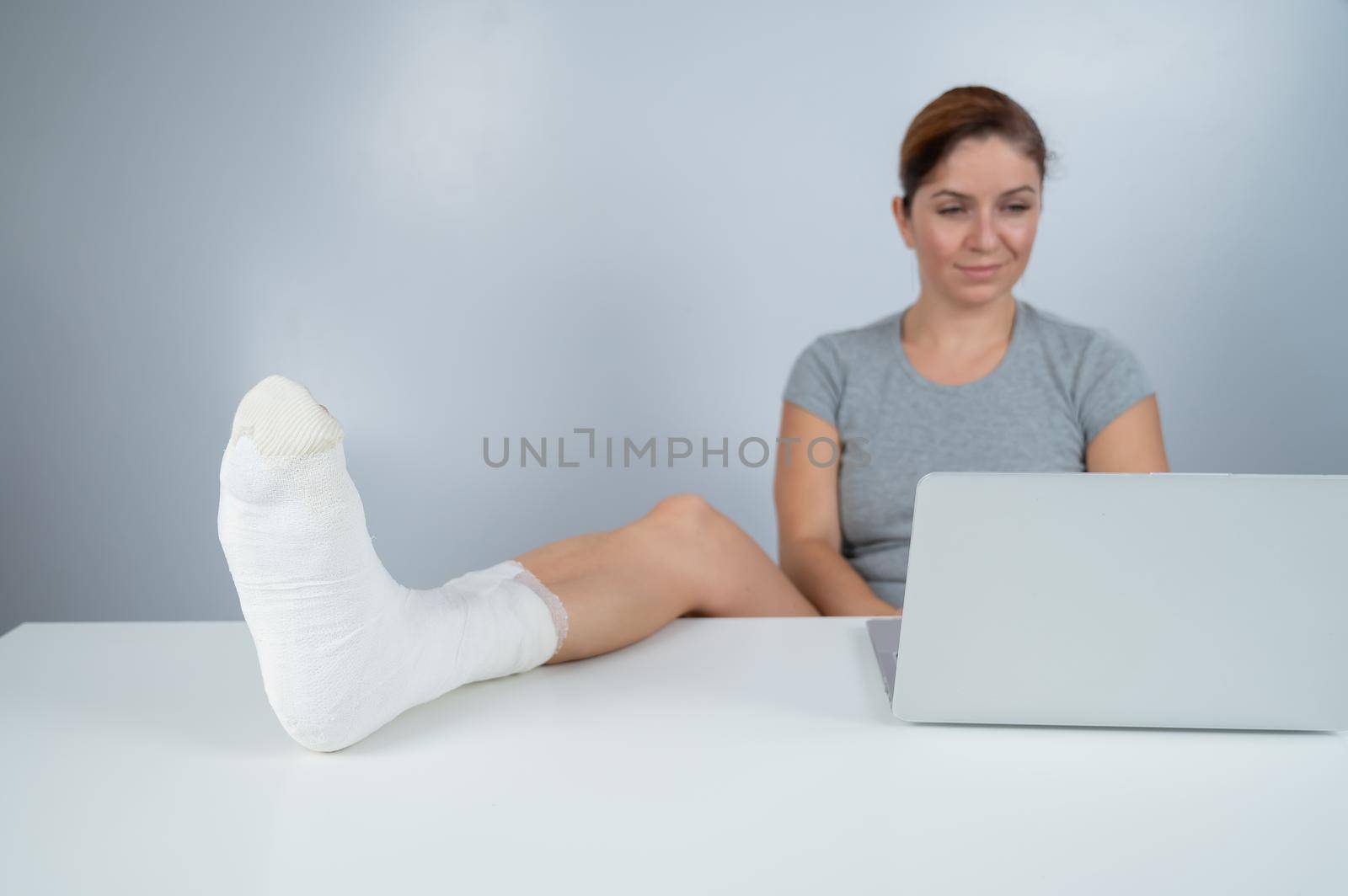 Caucasian woman lifted her leg with plaster to work desk and works on laptop on white background.