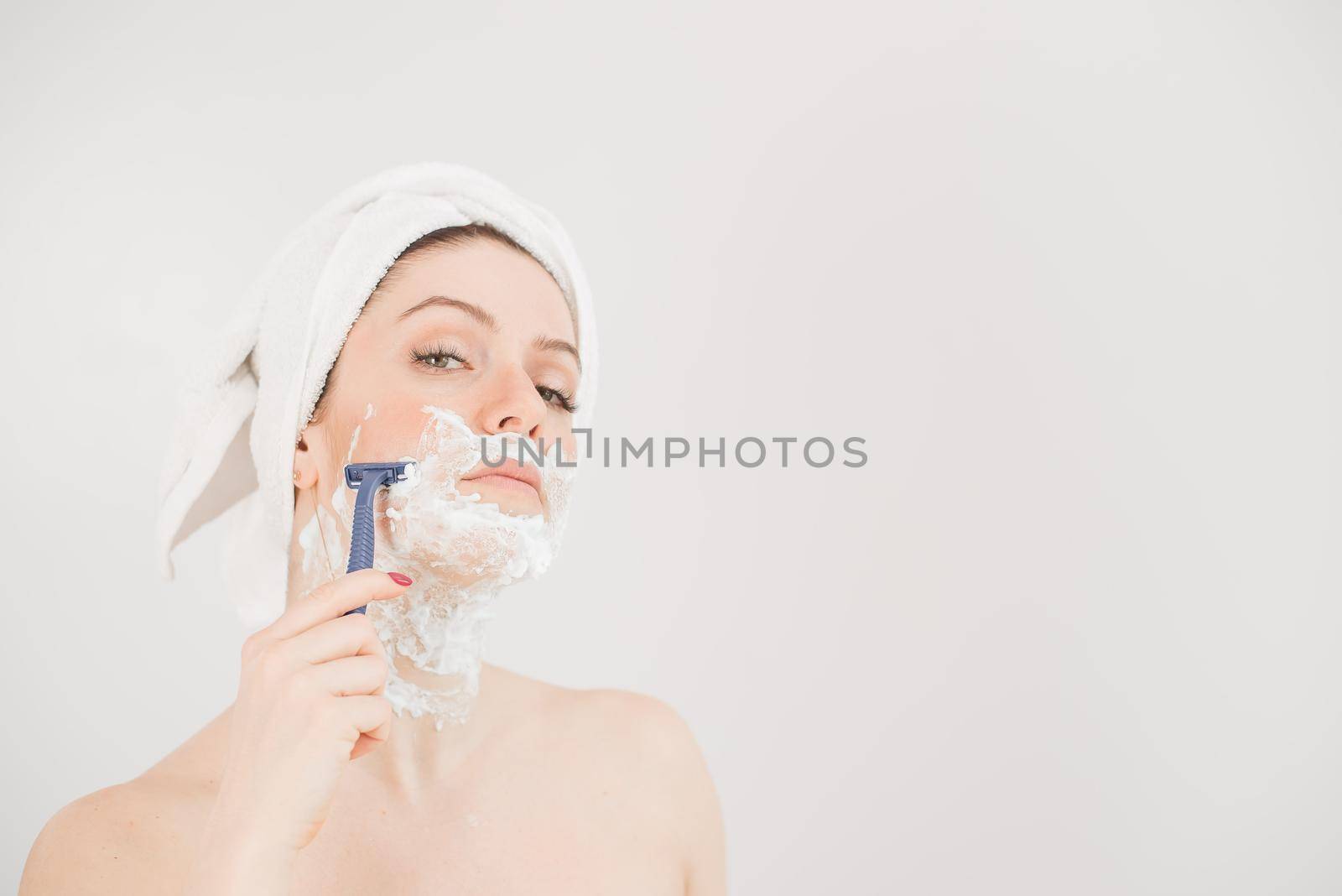 Cheerful caucasian woman with a towel on her head and shaving foam on her face holds a razor on a white background.