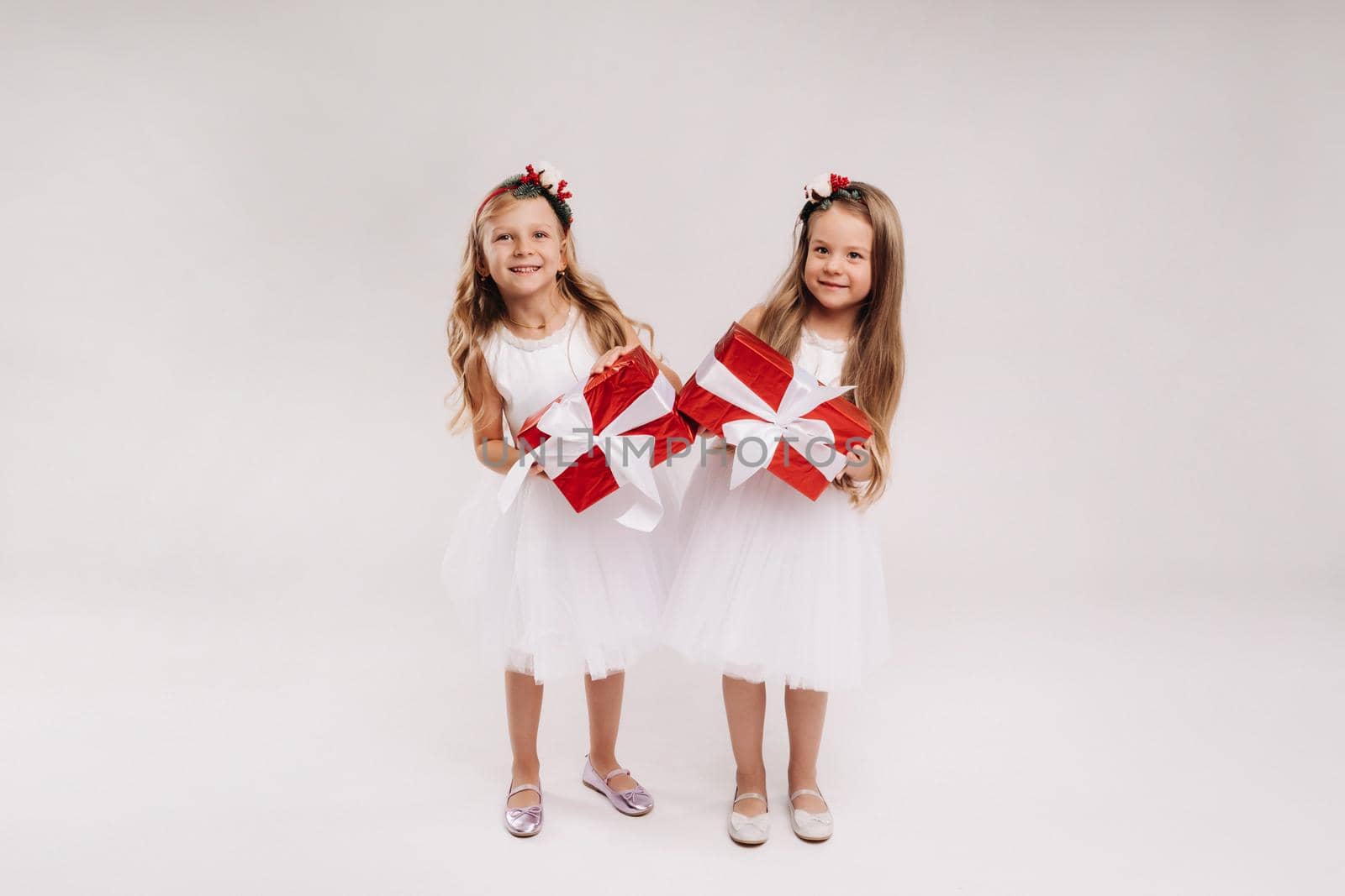 two little girls in white dresses with Christmas gifts on a white background smile by Lobachad