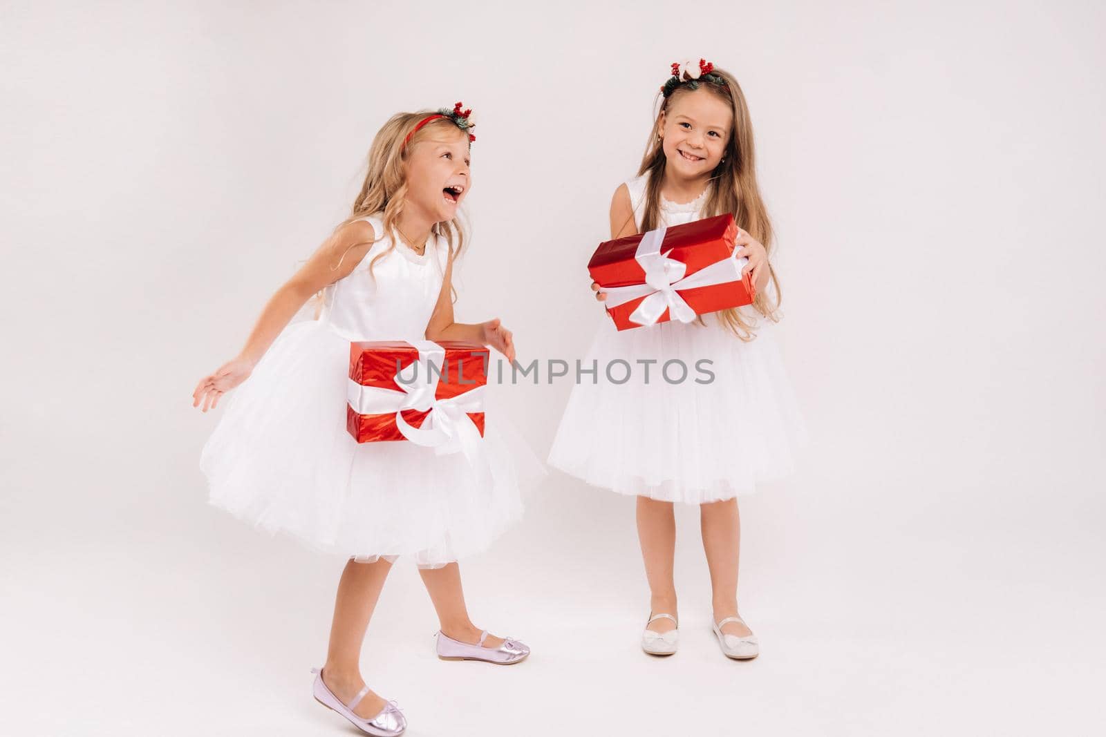 two little girls in white dresses with Christmas gifts on a white background smile.