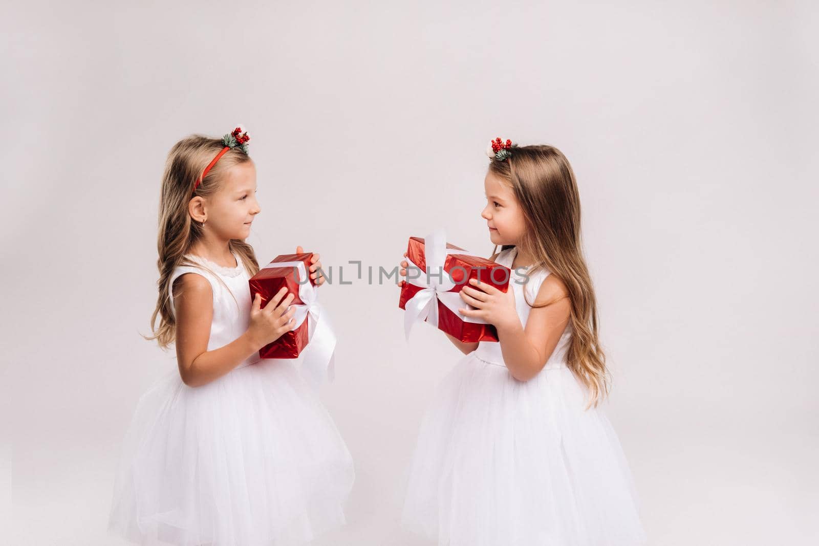 two little girls in white dresses with Christmas gifts on a white background smile by Lobachad