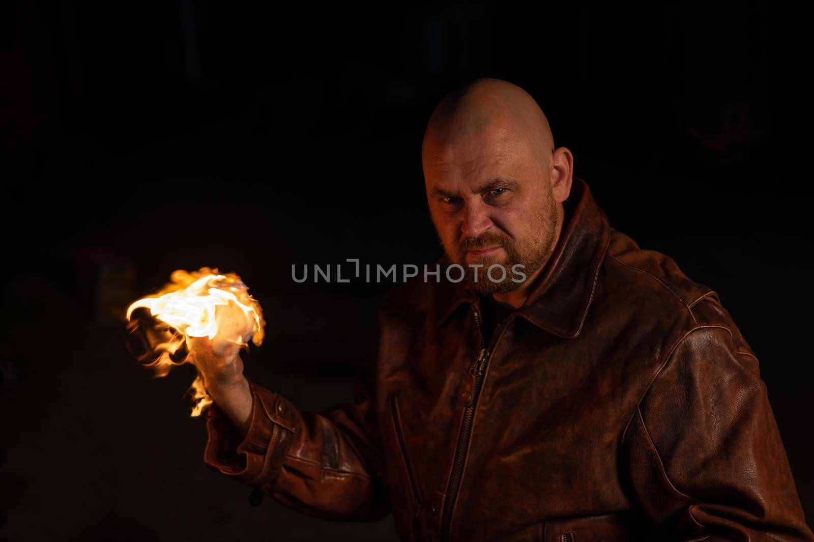 A bald man in a leather jacket holds a burning ball in his hand