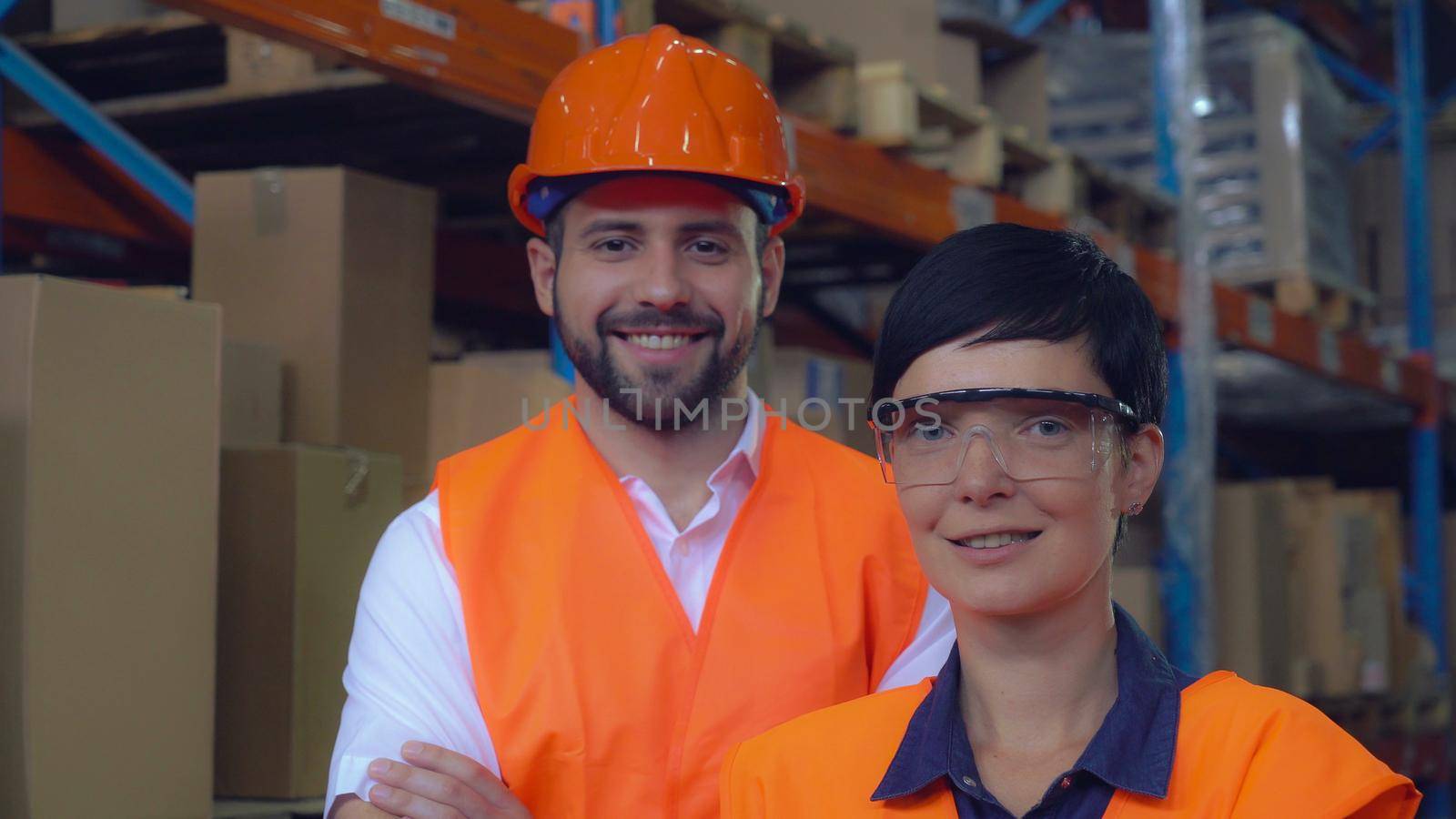 Portrait two workers at warehouse. Happy smiling co workers posing looking at the camera and smiling. Young professional woman and man wearing uniform high visibility orange hard hat ,vest