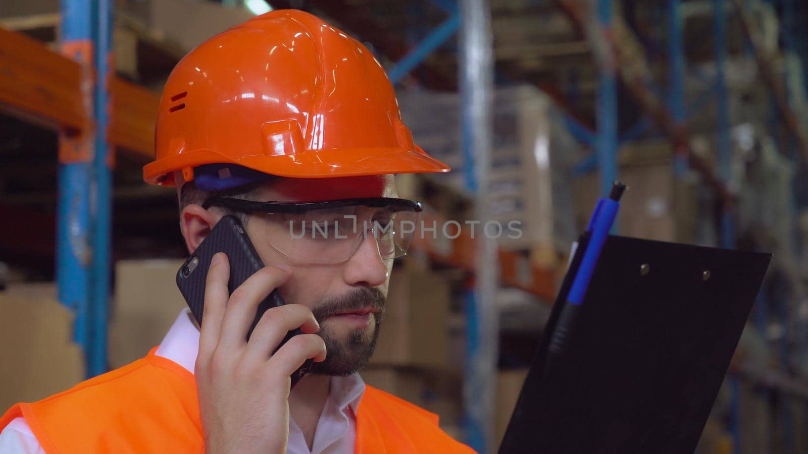 Portrait manager at work in warehouse. Handsome worker talking by smartphone discussing the logistics . Man with beard wearing hard hat and orange vest holding documents and checking delivery.