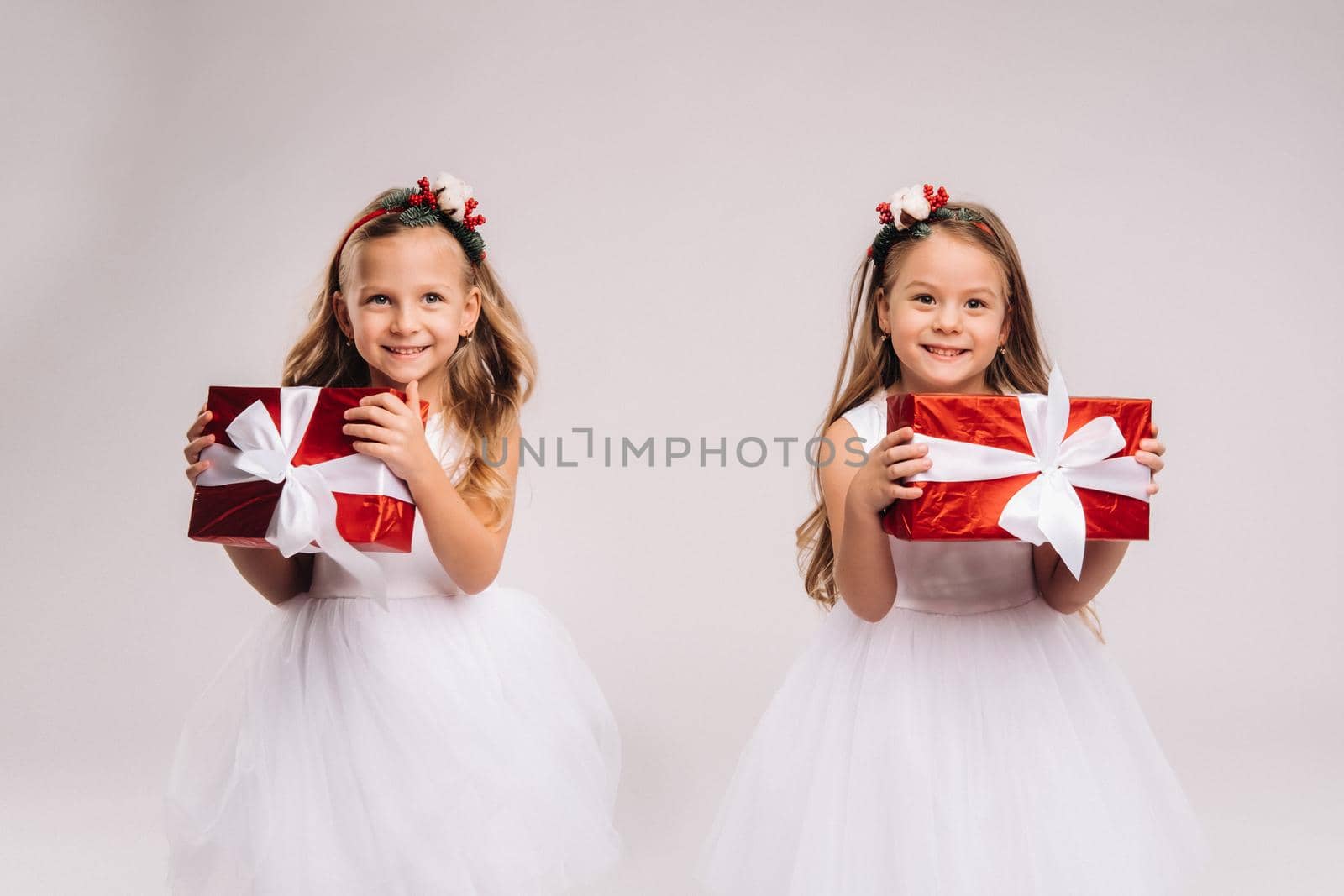 two little girls in white dresses with Christmas gifts on a white background smile.