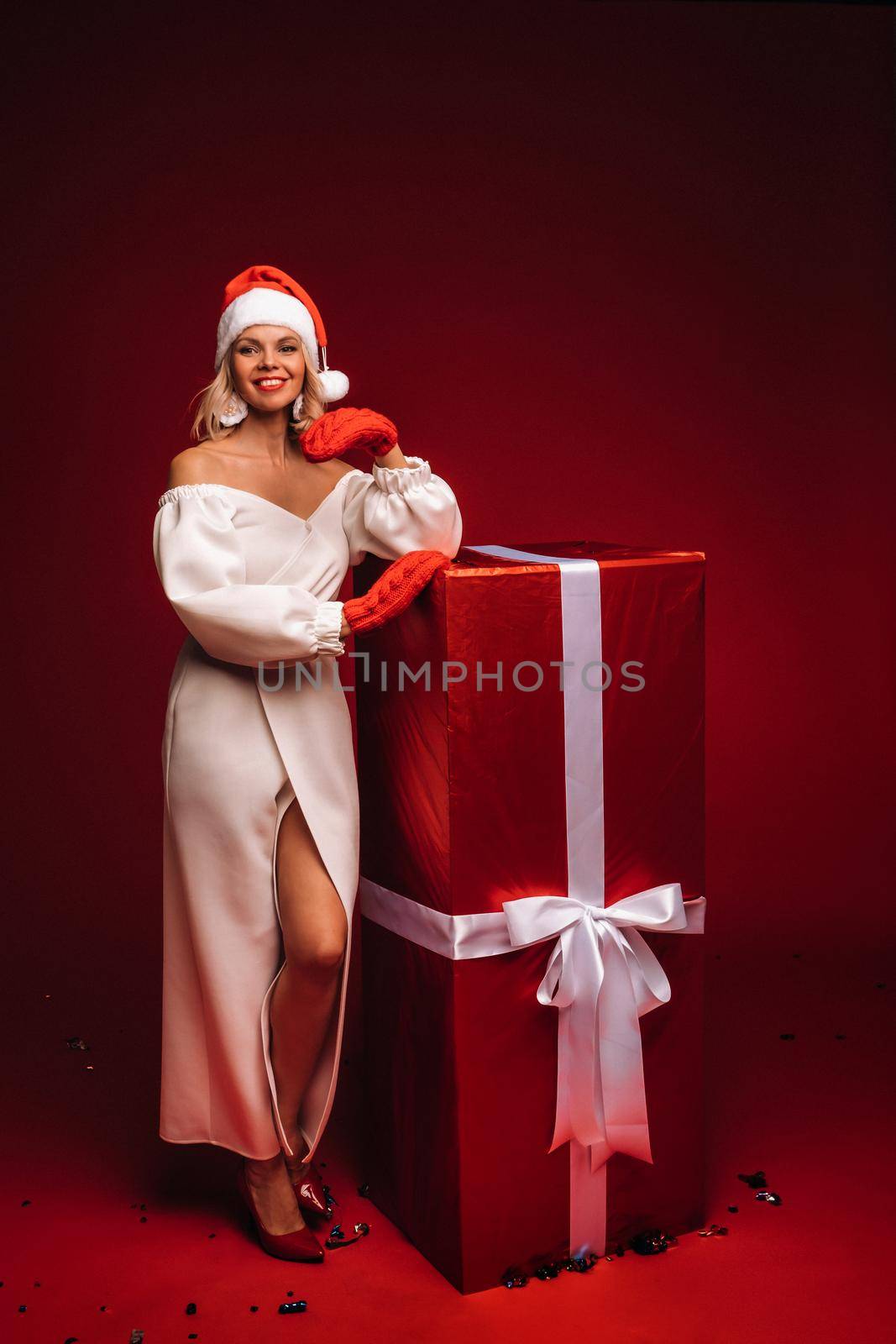 portrait of a smiling girl in a white dress and Santa hat with a huge Christmas gift on a red background.