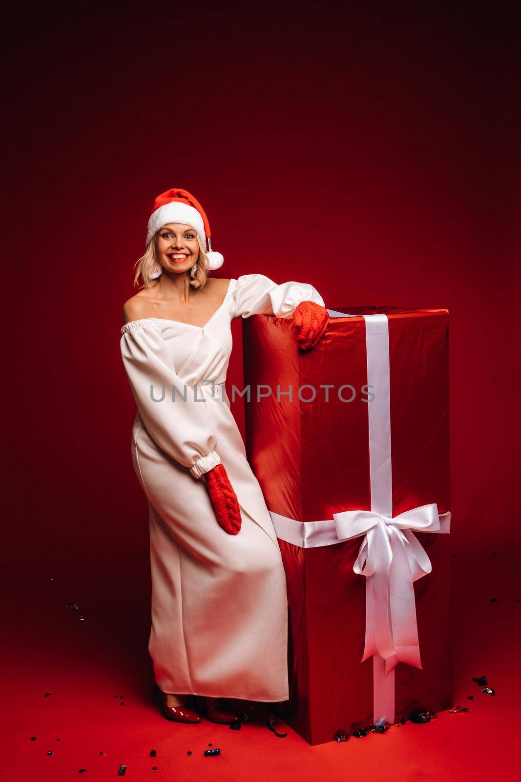 portrait of a smiling girl in a white dress and Santa hat with a huge Christmas gift on a red background.