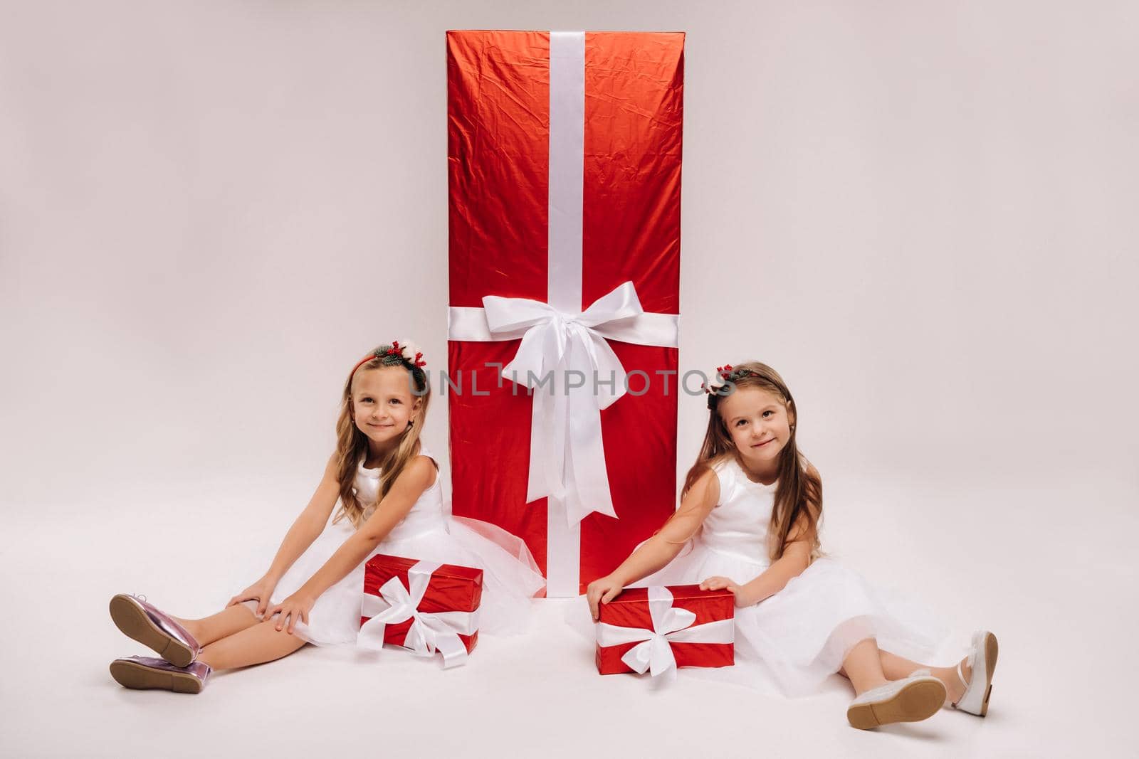 two little girls sit with Christmas gifts on a white background and a huge gift.
